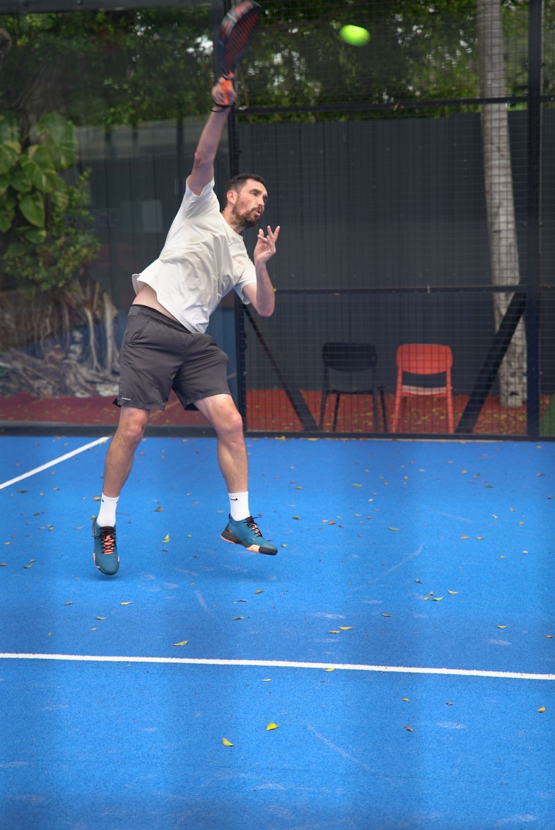 Tennis player serves ball on bright blue padel court during outdoor game on sunny day.