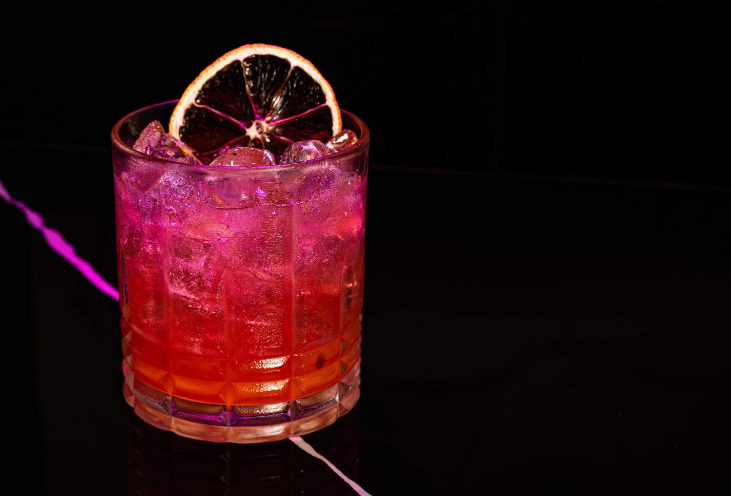 A bright pink cocktail garnished with a dried citrus wheel served in a rocks glass with ice against a dark background.
