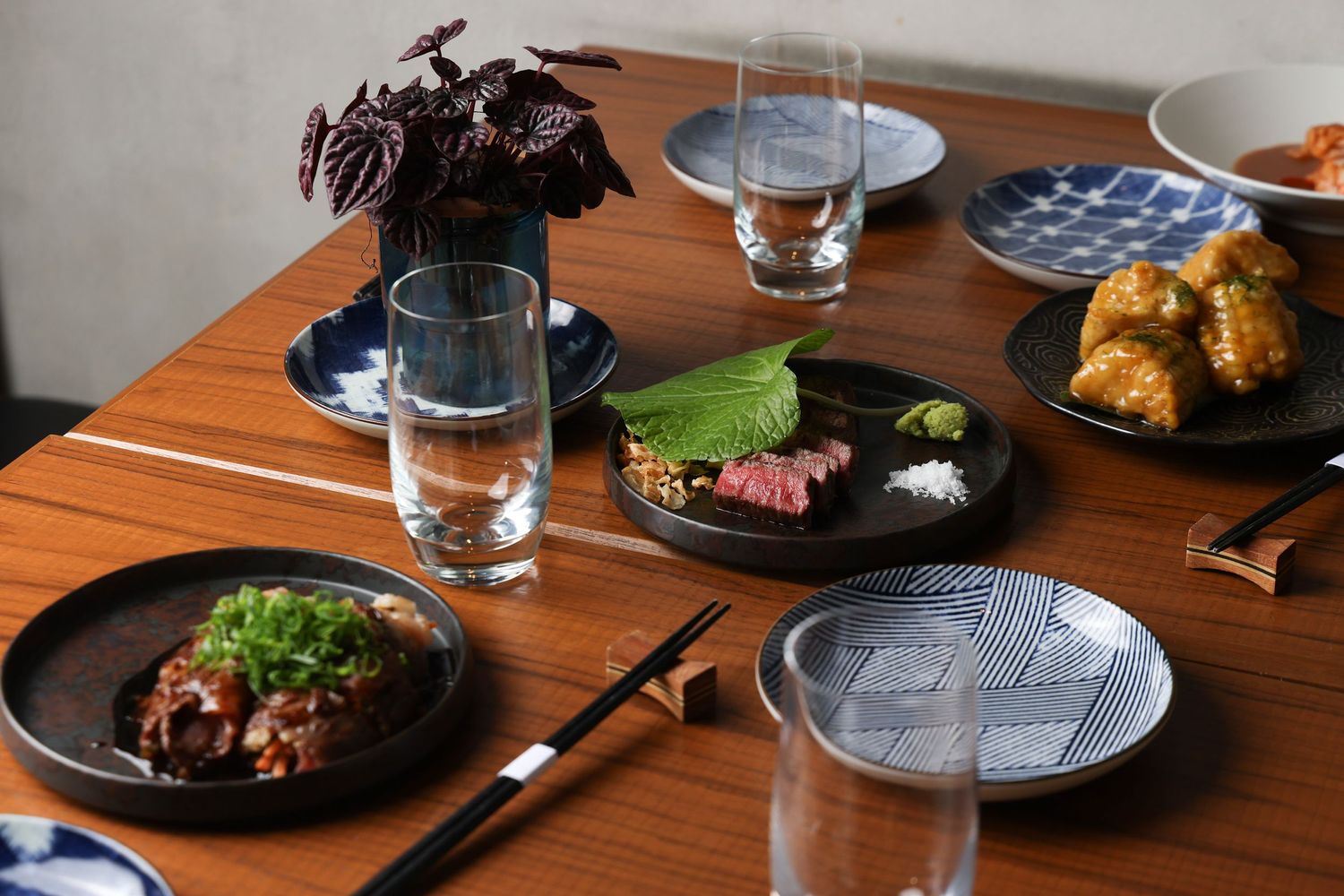 A modern dining table set with black plates of gourmet food, water glasses, and wooden tabletop in natural light.