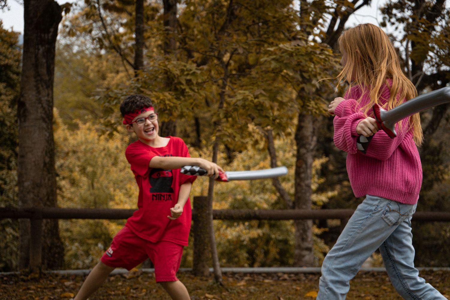 Two children playing with toy swords on an autumn day near a wooden fence.