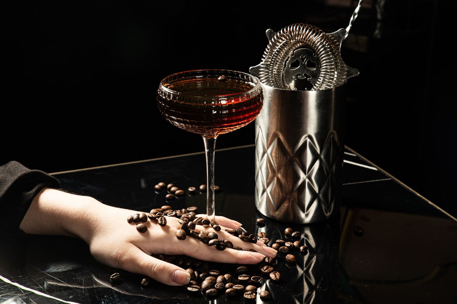 Elegant cocktail setup with metal shaker, coffee beans, and dark red drink in coupe glass on black background.