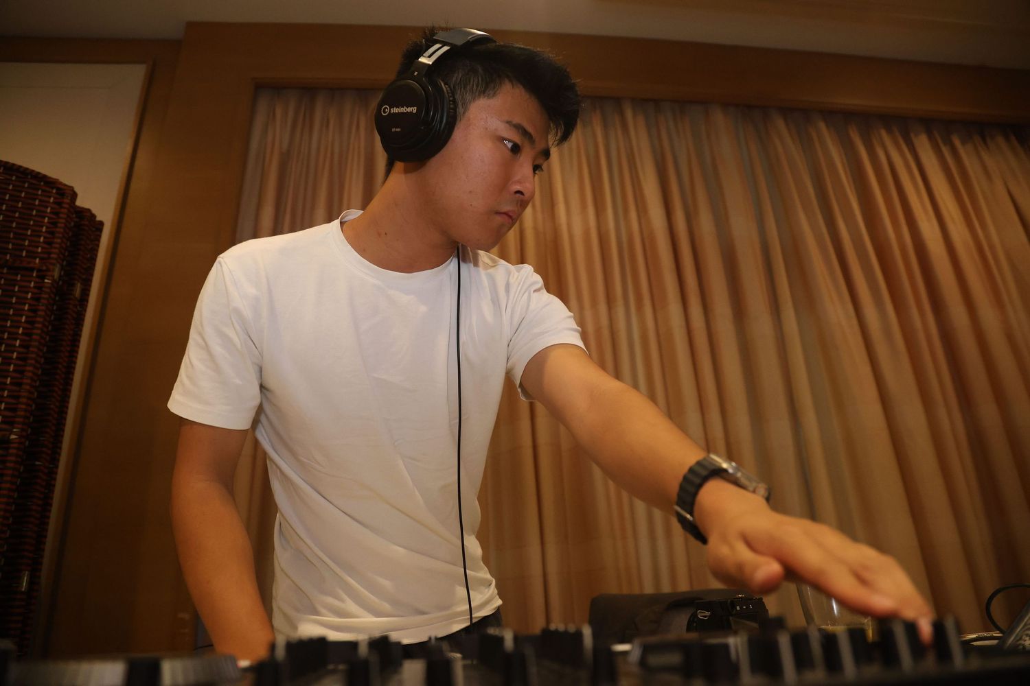 DJ wearing headphones and a white t-shirt performs at a mixing console in a dimly lit room.