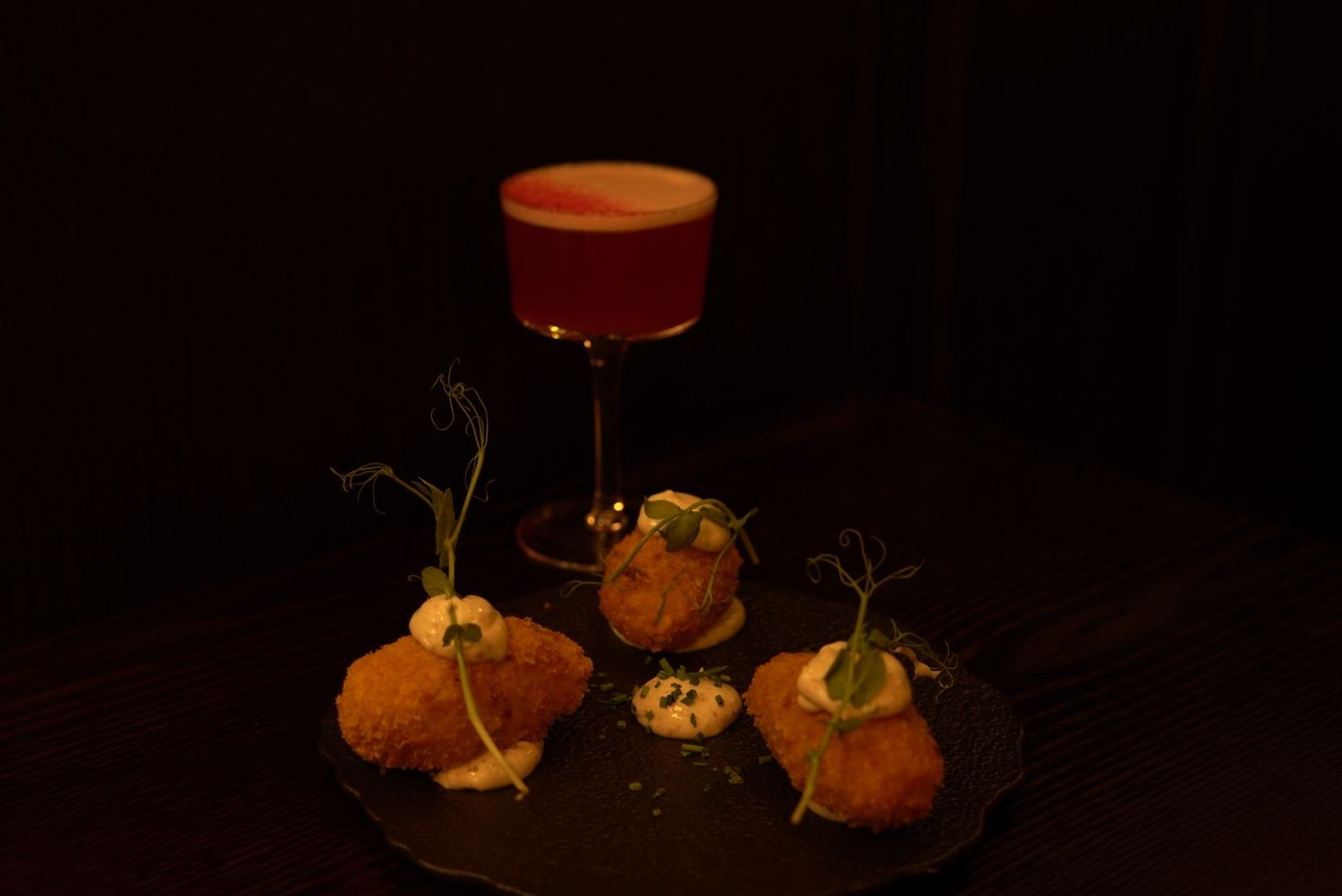 Red cocktail and orange appetizers plated on dark surface with dramatic lighting.