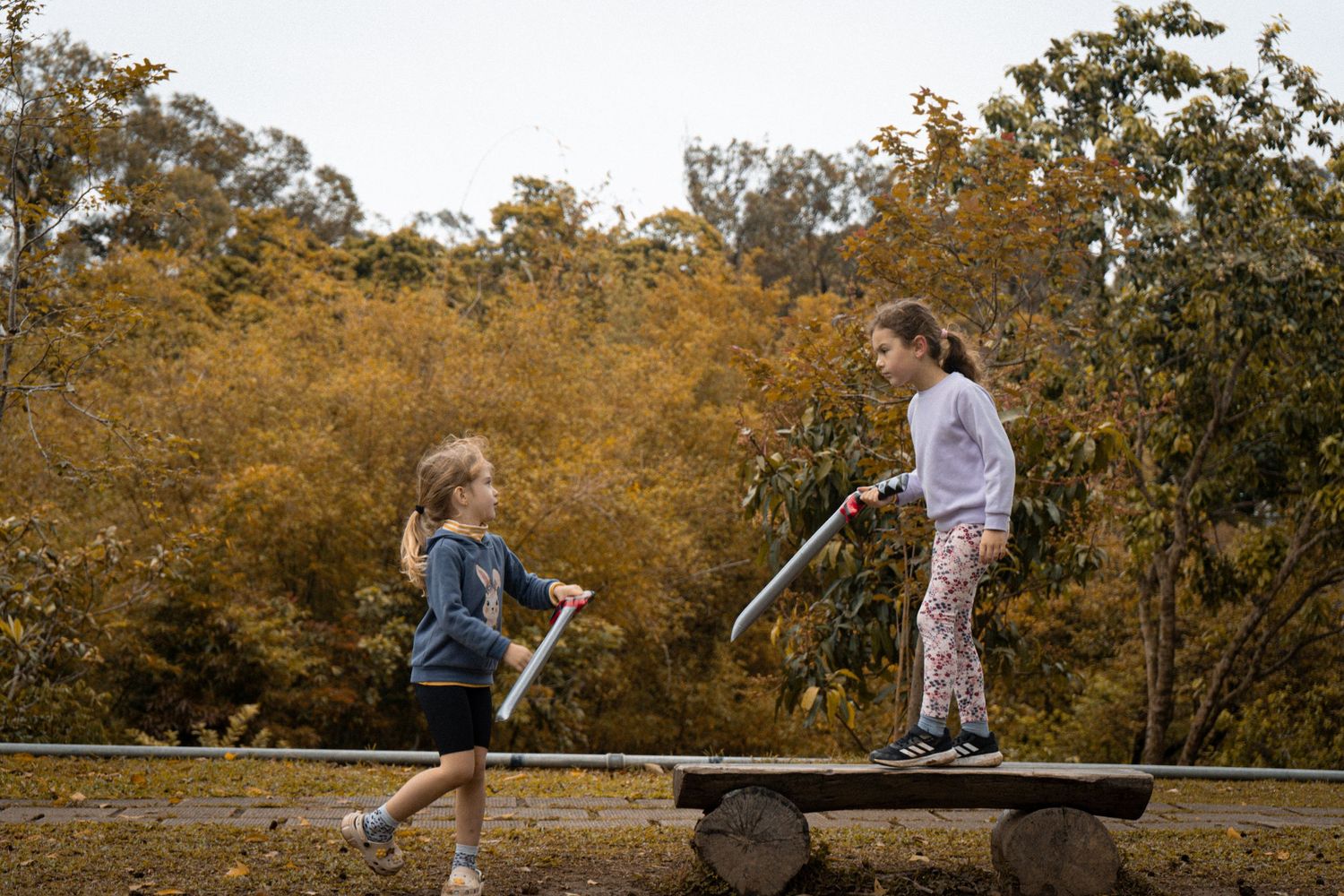 Two children playing with toy swords while balancing on a wooden board outdoors.