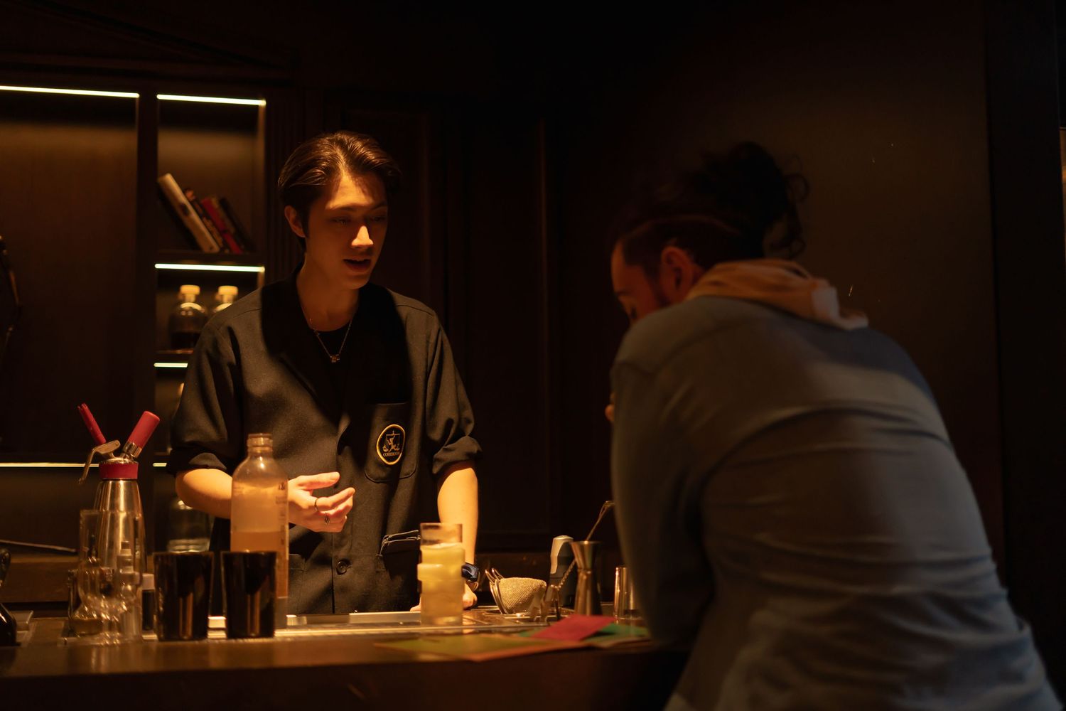Two figures sit at a dimly lit bar counter with bottles and glasses visible in the atmospheric lighting.