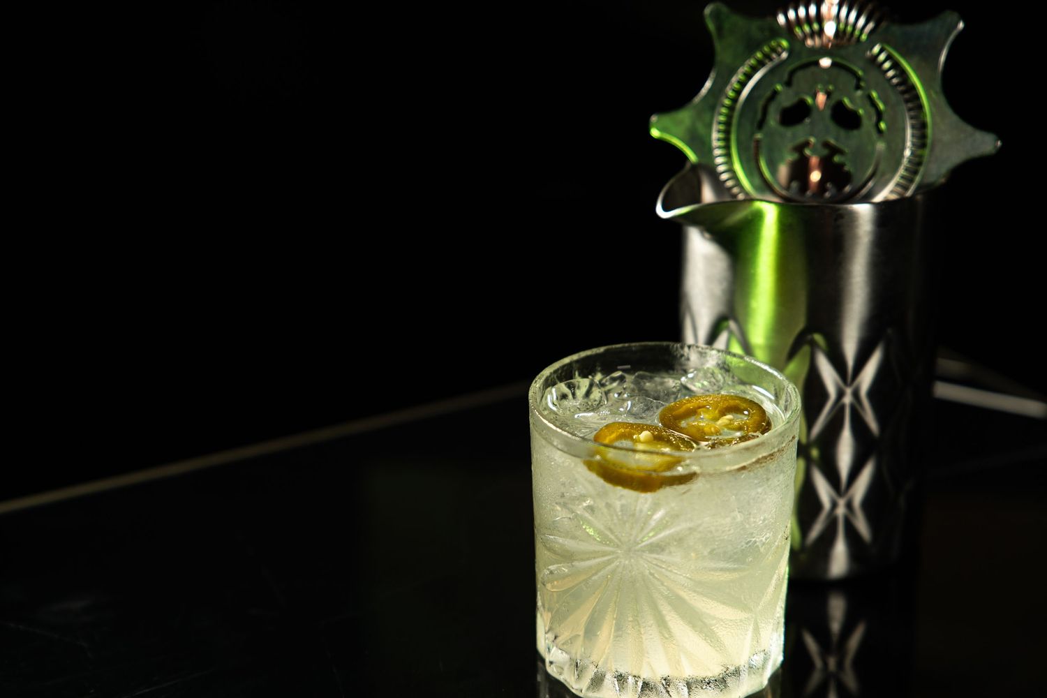 An elegant cocktail in a cut crystal glass with a decorative metal garnish holder on a black surface.