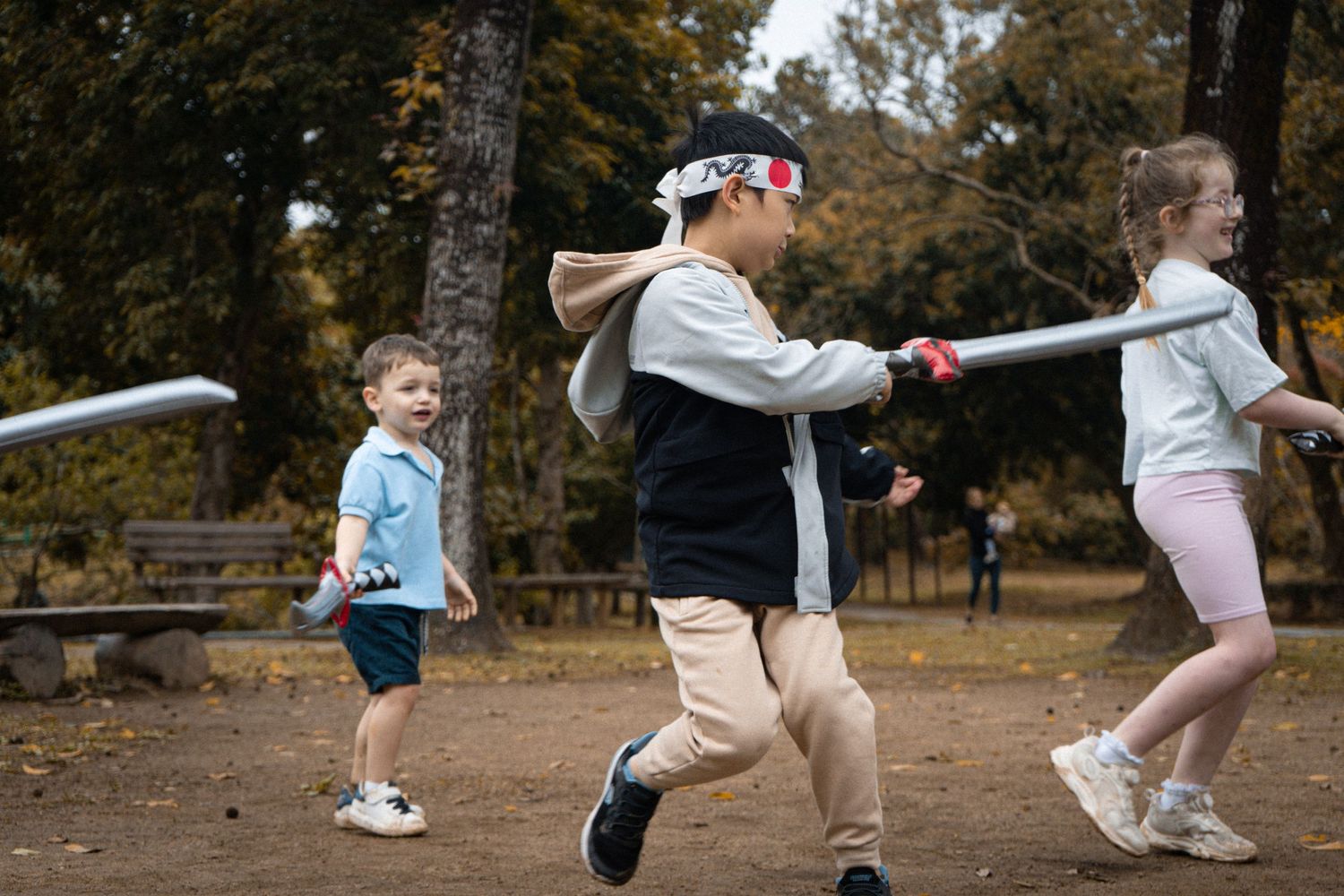 Children engaged in playful sword fighting game in a park during fall season.
