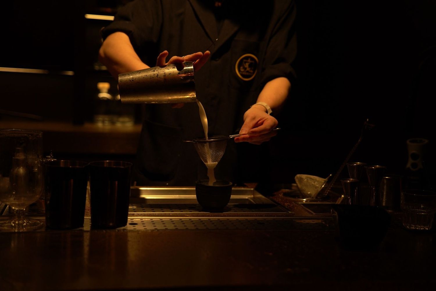 A bartender pours a drink in a dimly lit bar setting with dark ambient lighting.