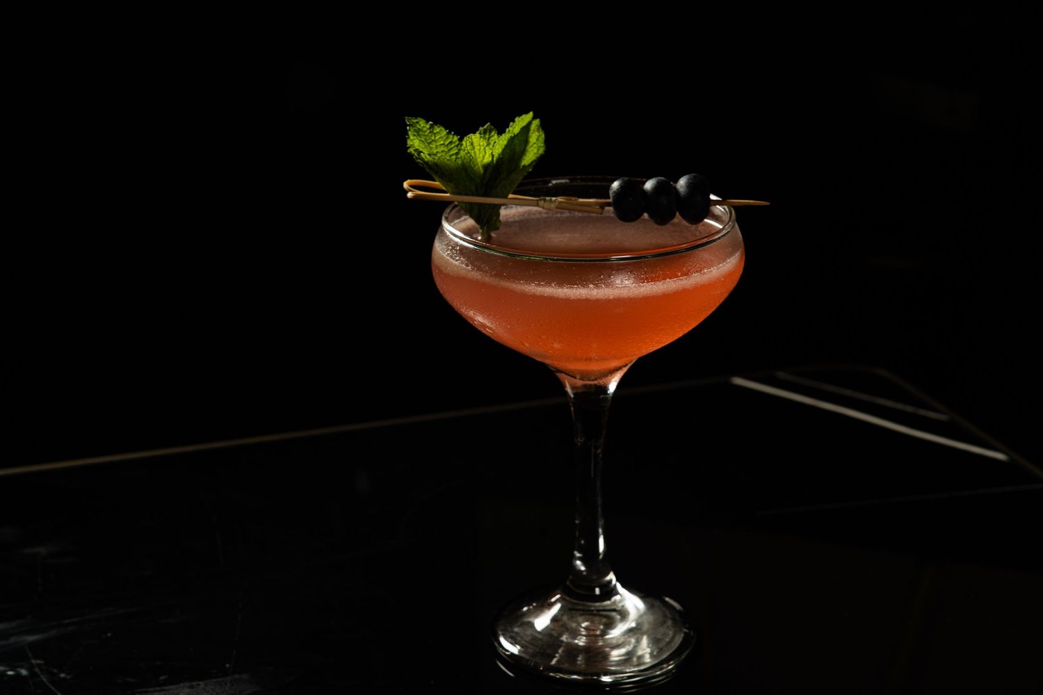 Pink cocktail in coupe glass garnished with mint leaves and berries on dark surface.