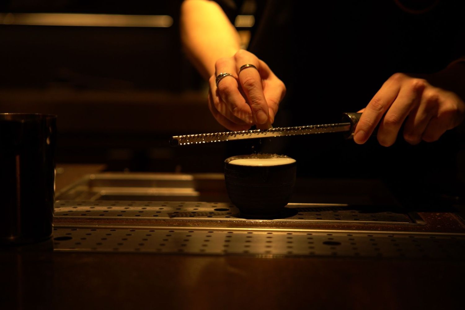 Someone grating spices or cheese onto a dark bowl in moody lighting at a restaurant or kitchen counter.