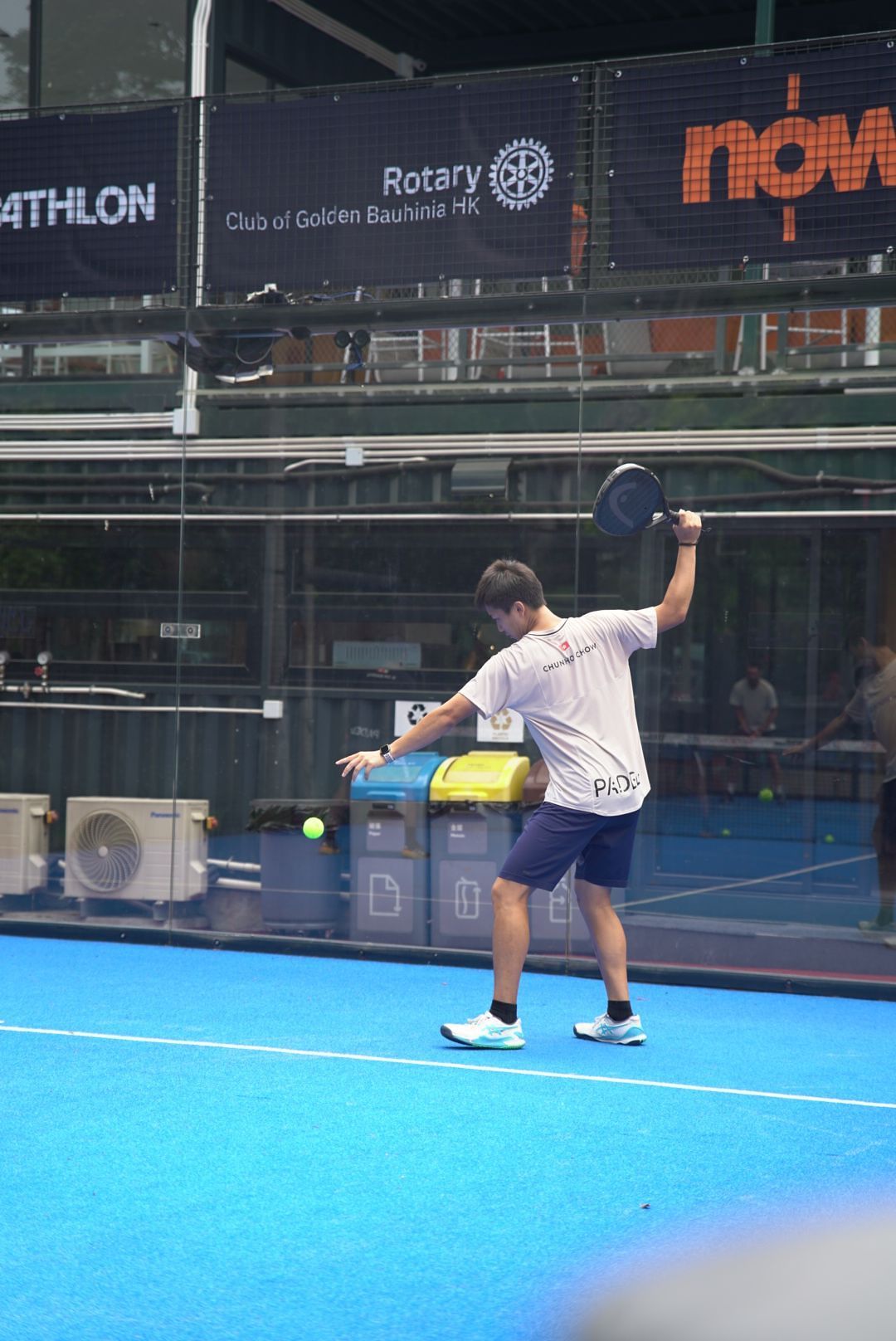 Player executing a backhand volley on an indoor blue padel court with Rotary Club signage visible.