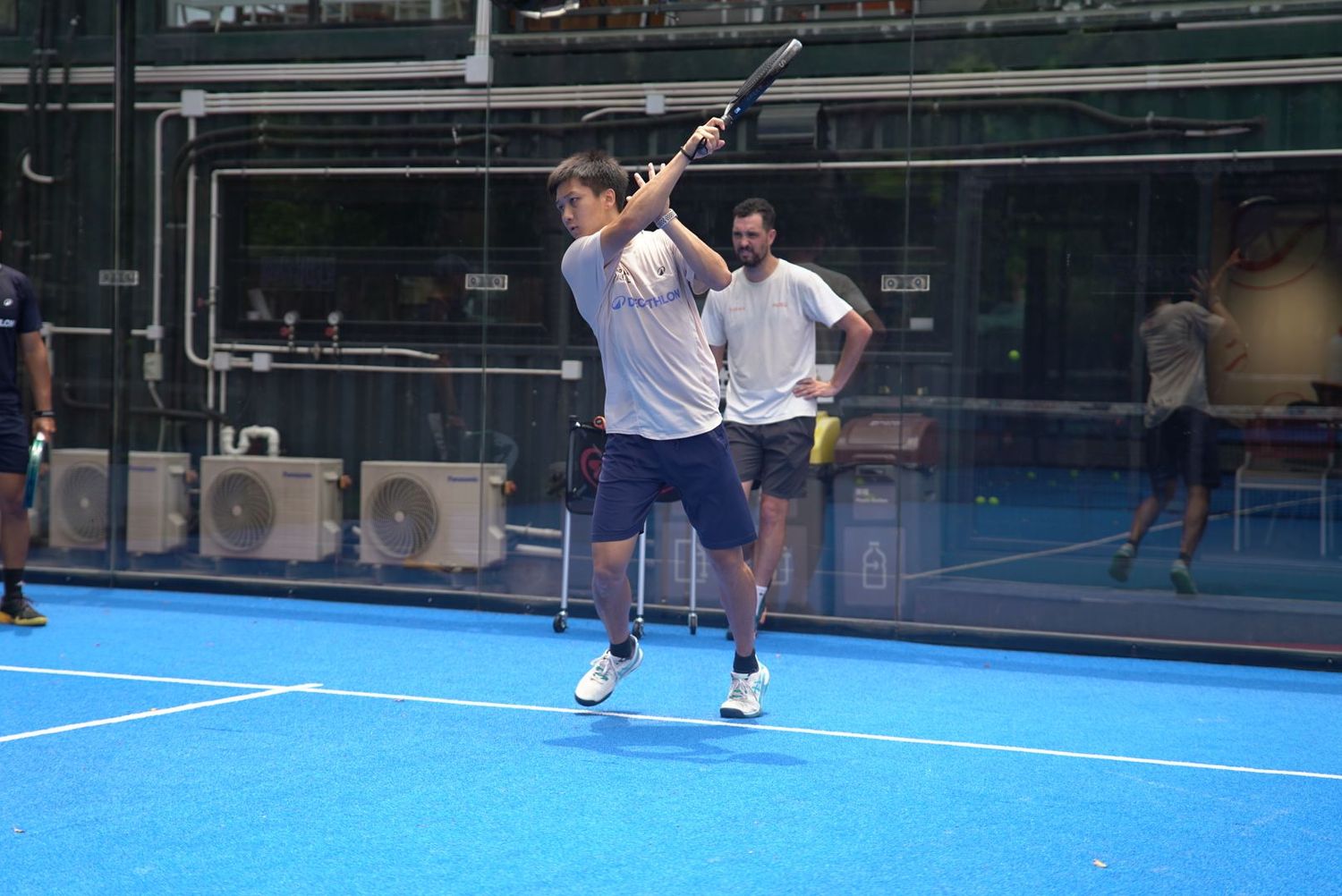 Tennis player practicing serves on an outdoor blue court with glass walls and training equipment visible.