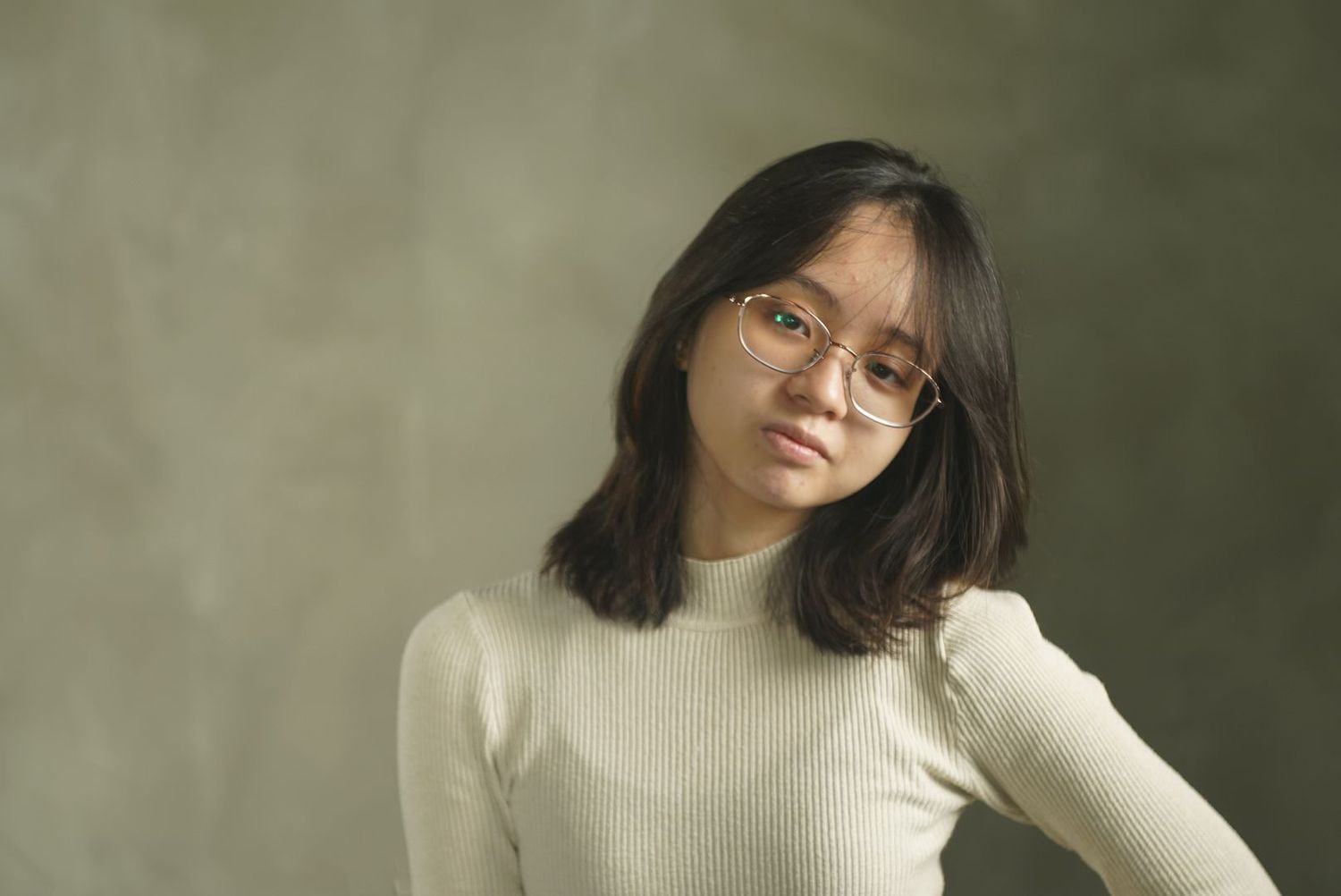 A person wearing glasses and a white sweater poses against a neutral backdrop in a moody portrait.