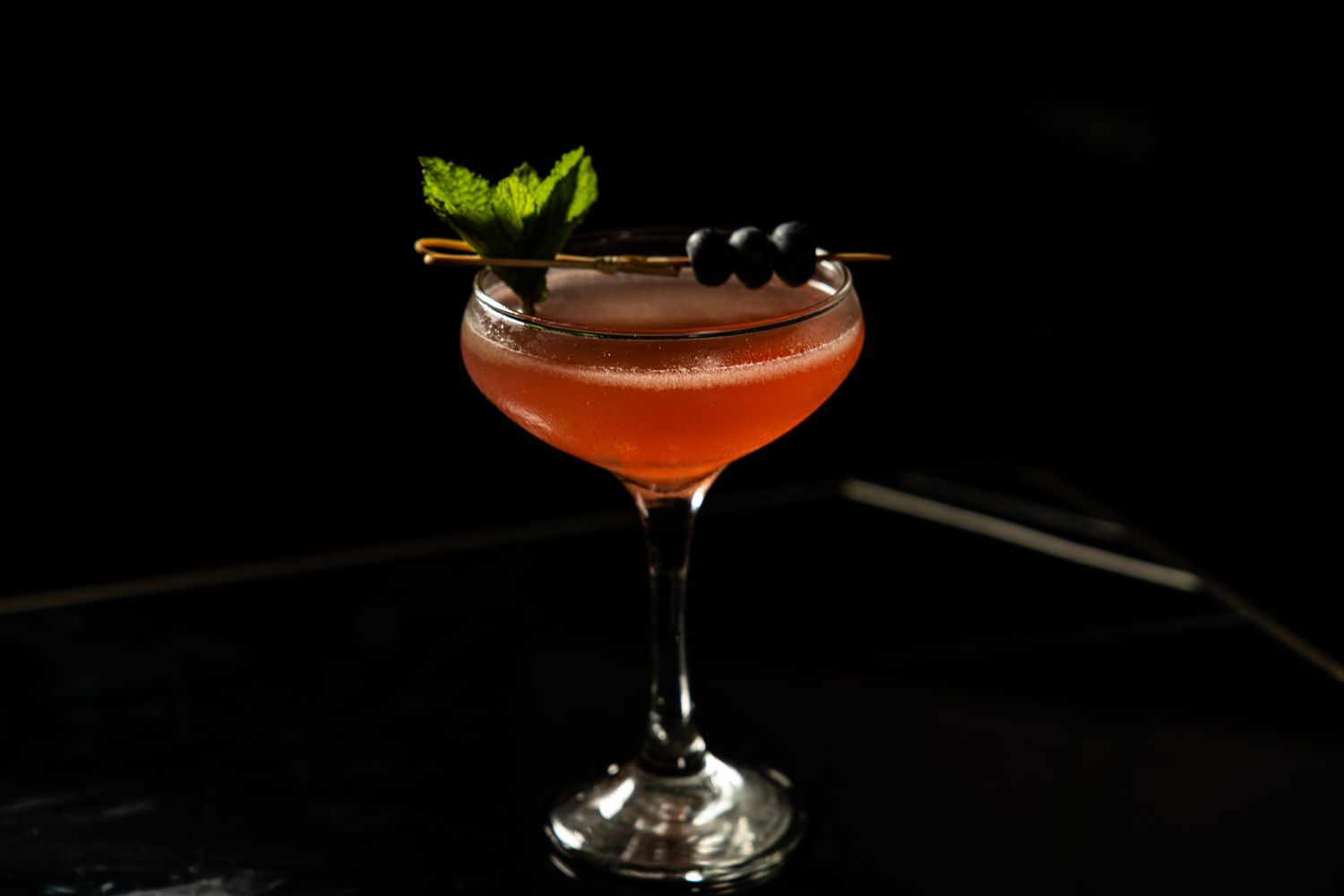 A vibrant red cocktail garnished with fresh mint leaves and dark berries in a classic coupe glass.