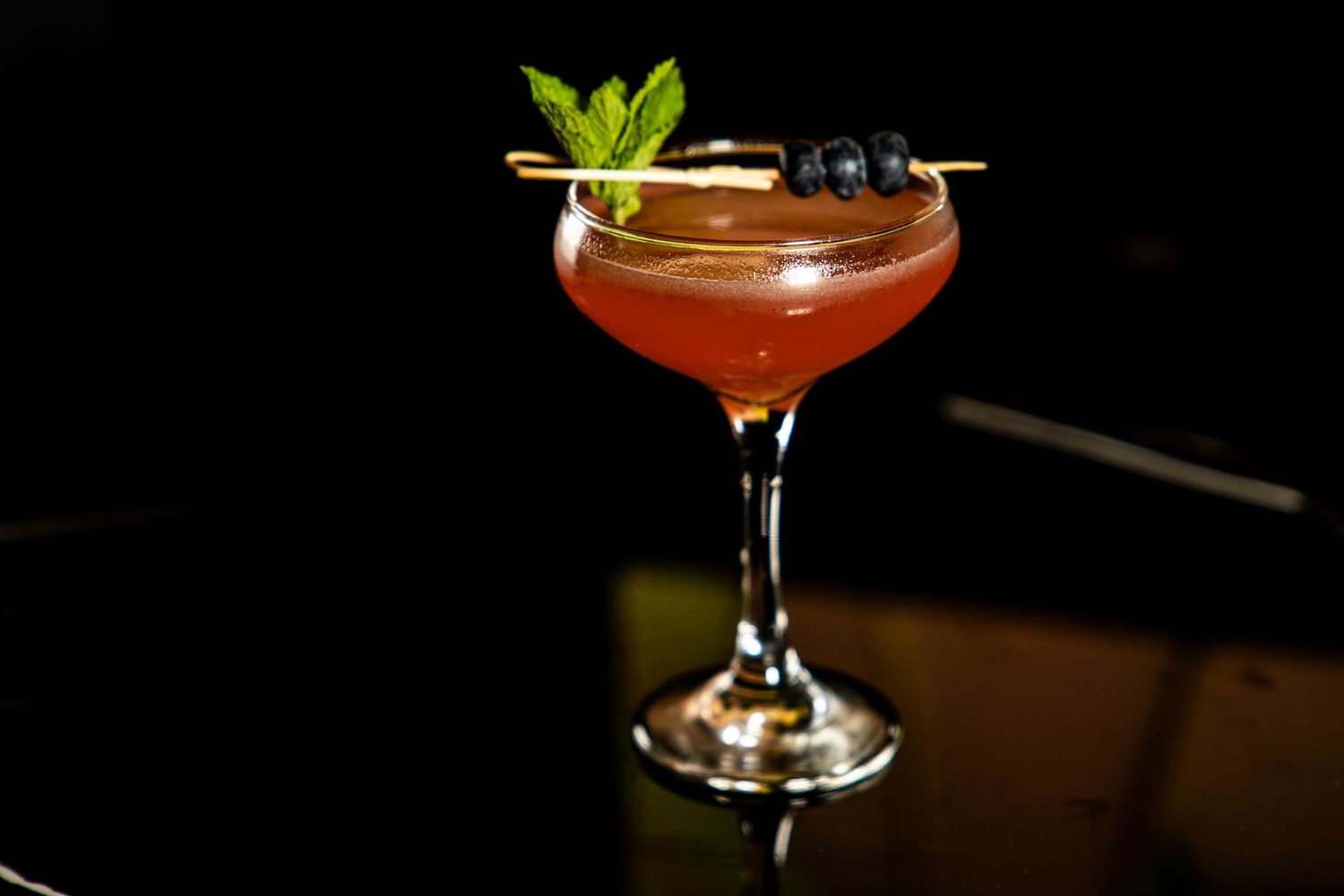 An elegant red cocktail in a coupe glass garnished with fresh mint leaves and dark berries against a black background.