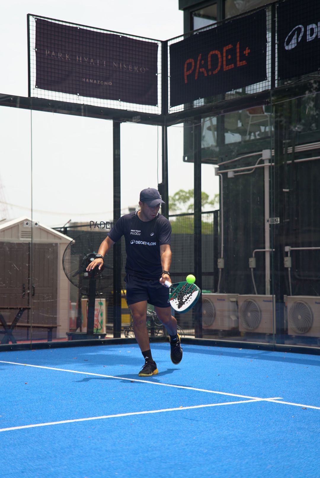 Padel player in dark athletic wear hits a backhand shot on blue court during match at indoor facility.