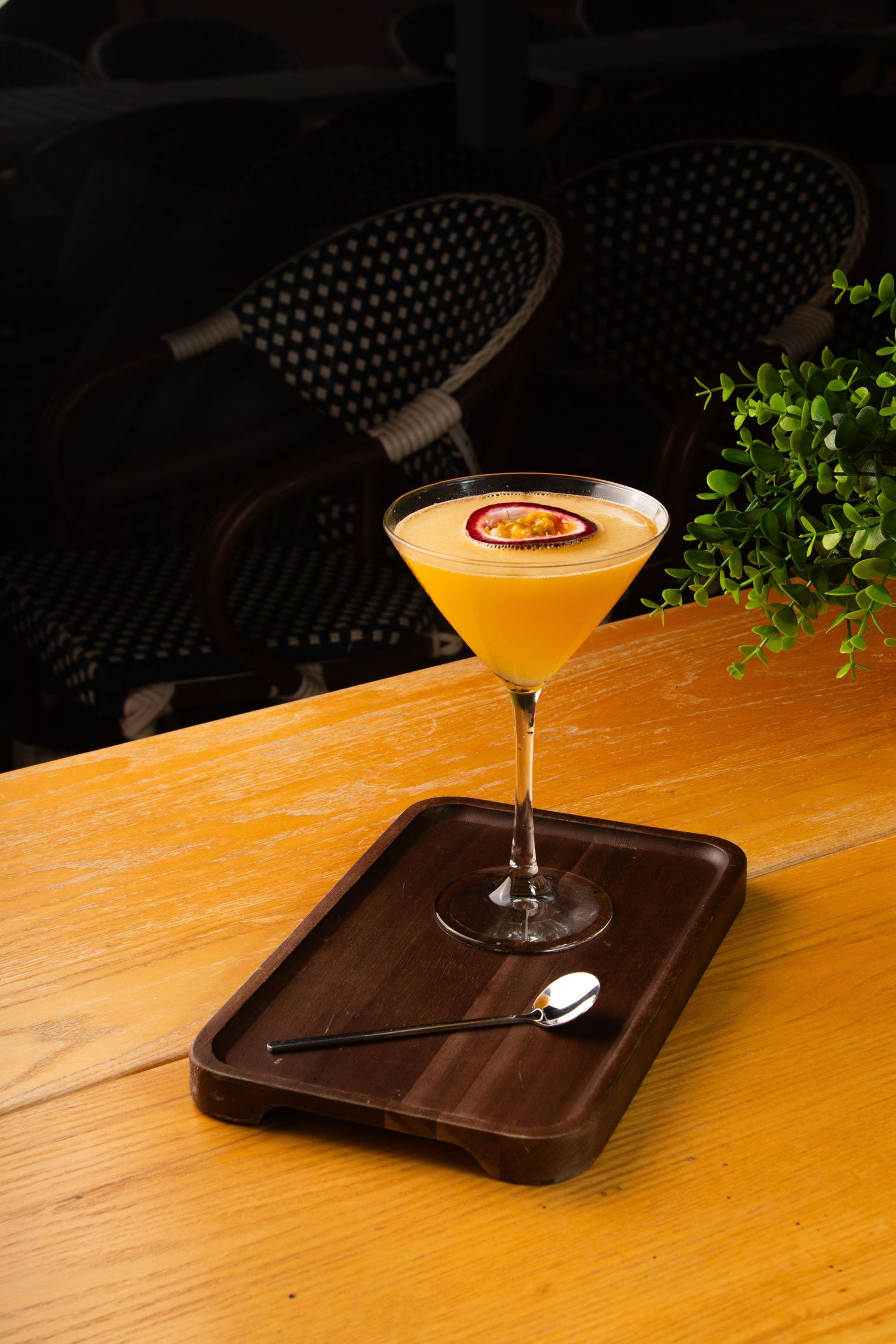 A yellow martini cocktail served in a coupe glass on a wooden tray with a spoon and decorative plant garnish.