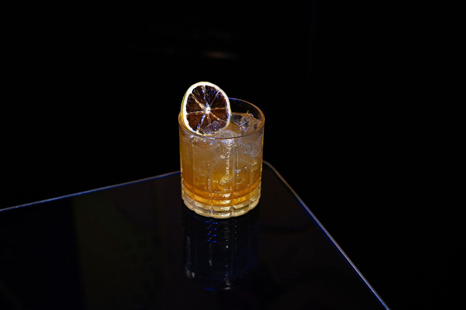 Amber cocktail with dehydrated orange garnish on reflective black surface.