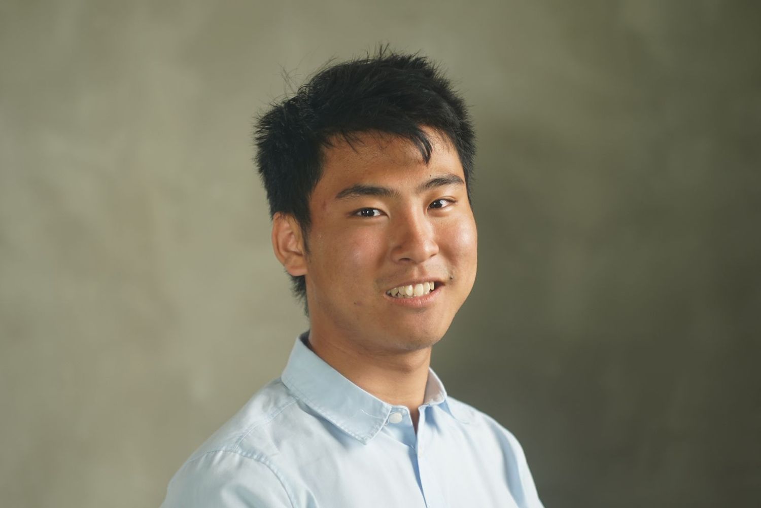 A young professional in a white button-down shirt smiles warmly against a blurred olive-colored background.