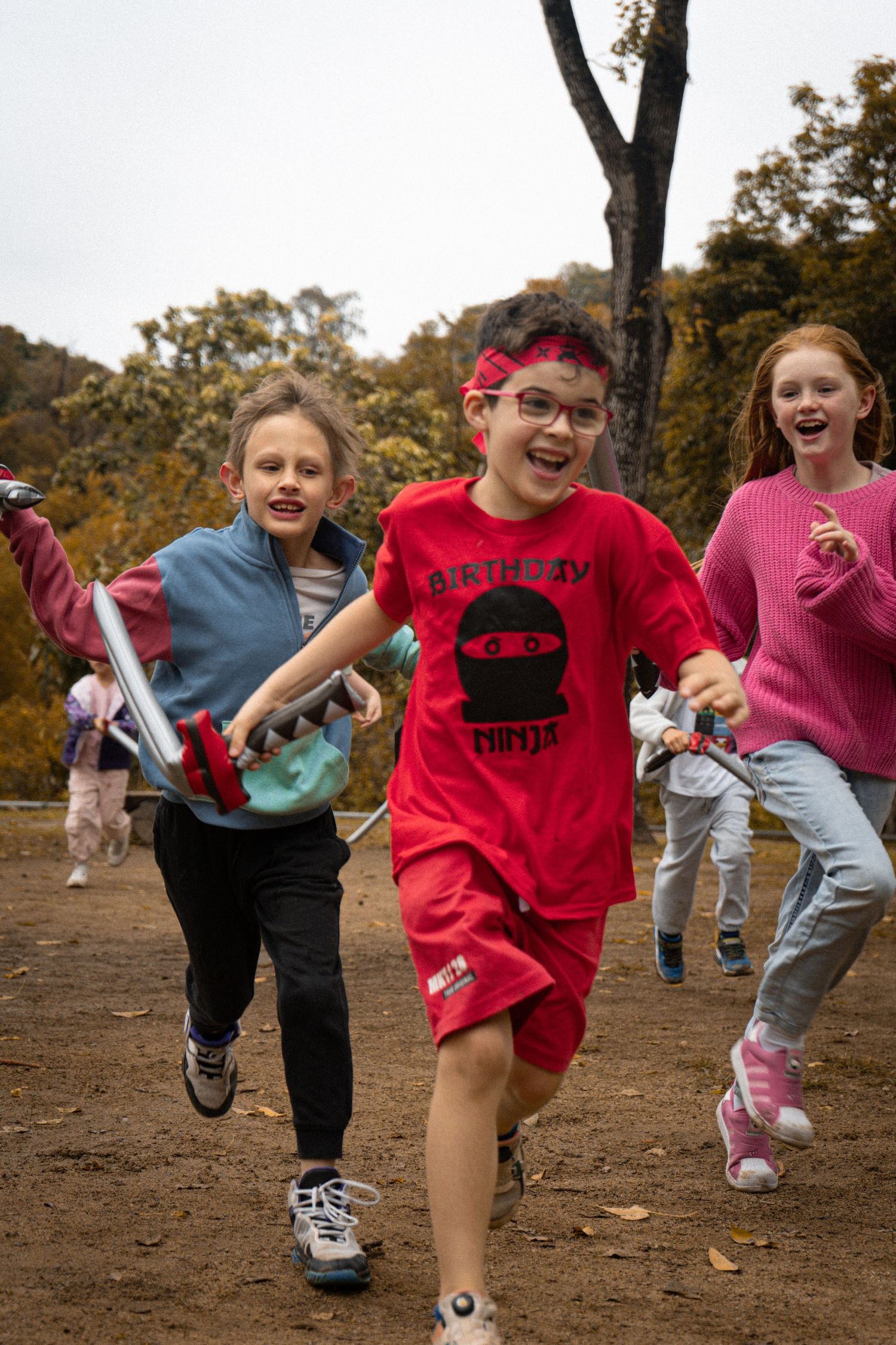 Children running with toy swords and playing games in an autumn park setting.