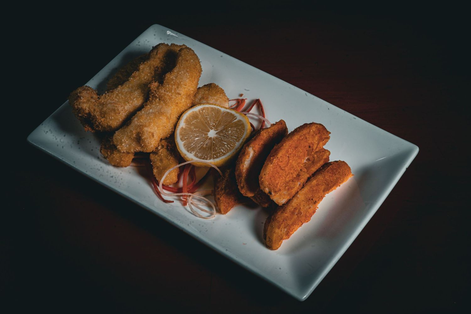 Breaded fried items served with lemon slice and red garnish on rectangular white plate.