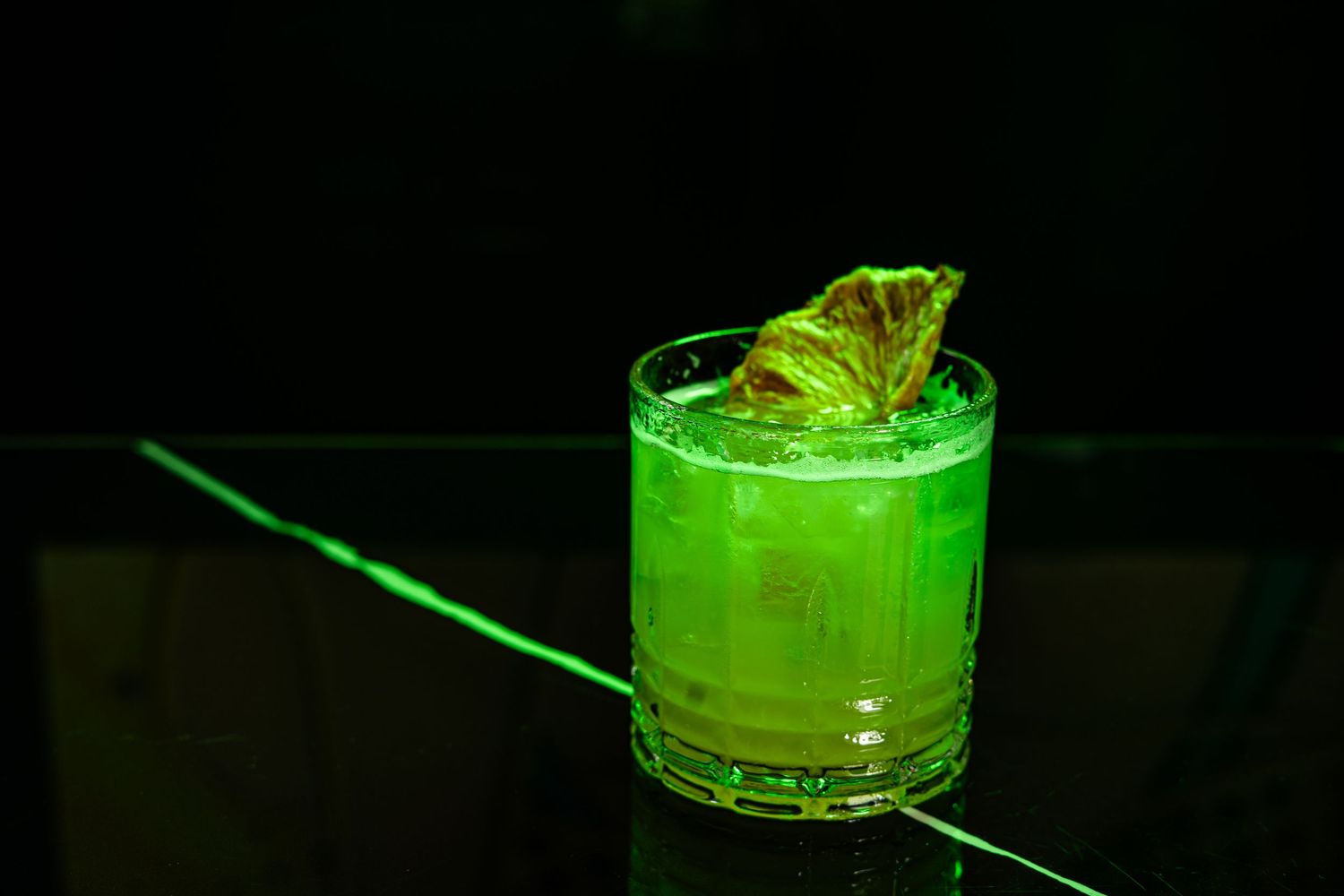 A vibrant green cocktail garnished with mint leaf glows in a rocks glass against a dark background.