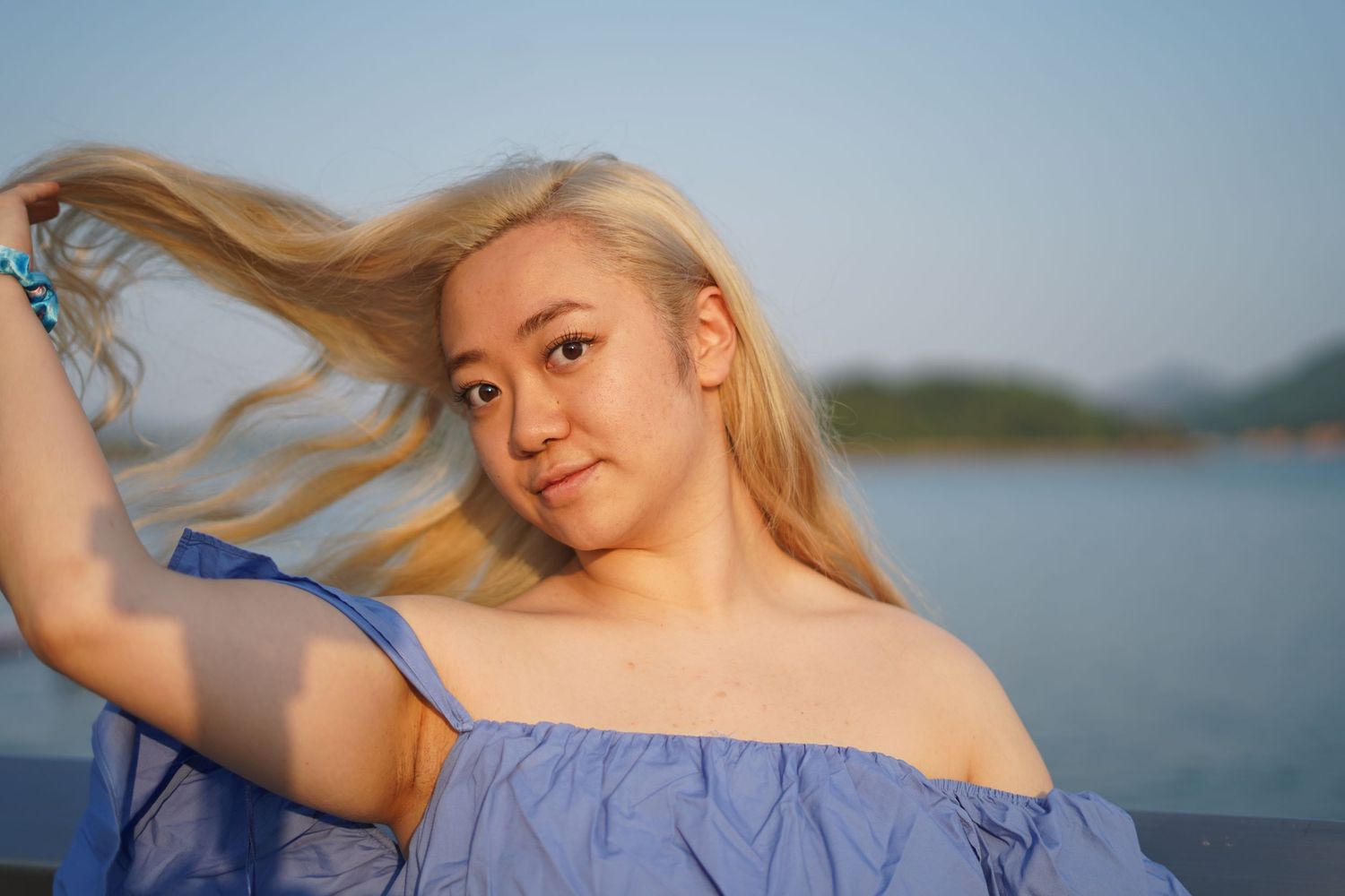 A person in an off-shoulder blue dress poses near water during a sunset photoshoot.