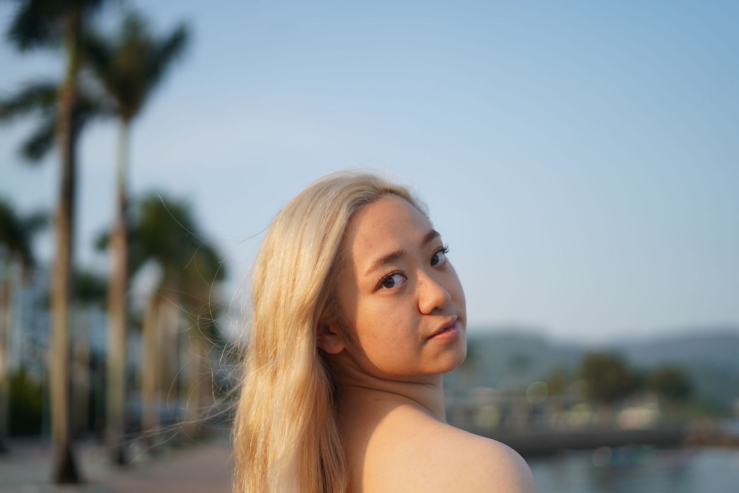 A person with long blonde hair poses near palm trees at sunset with a mountain view in the background.