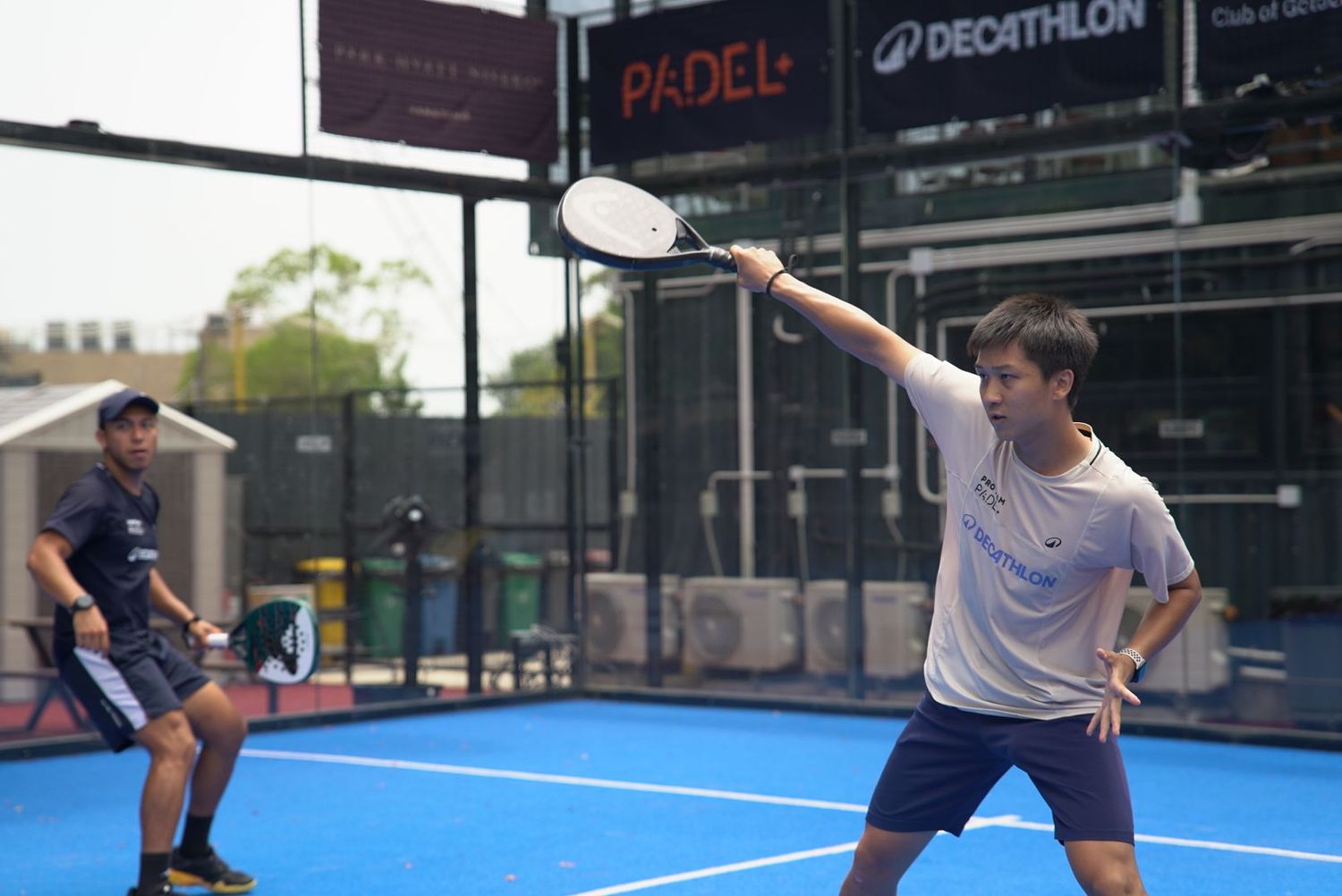 Two players compete in an outdoor padel match on a blue court with protective glass walls surrounding the playing area.