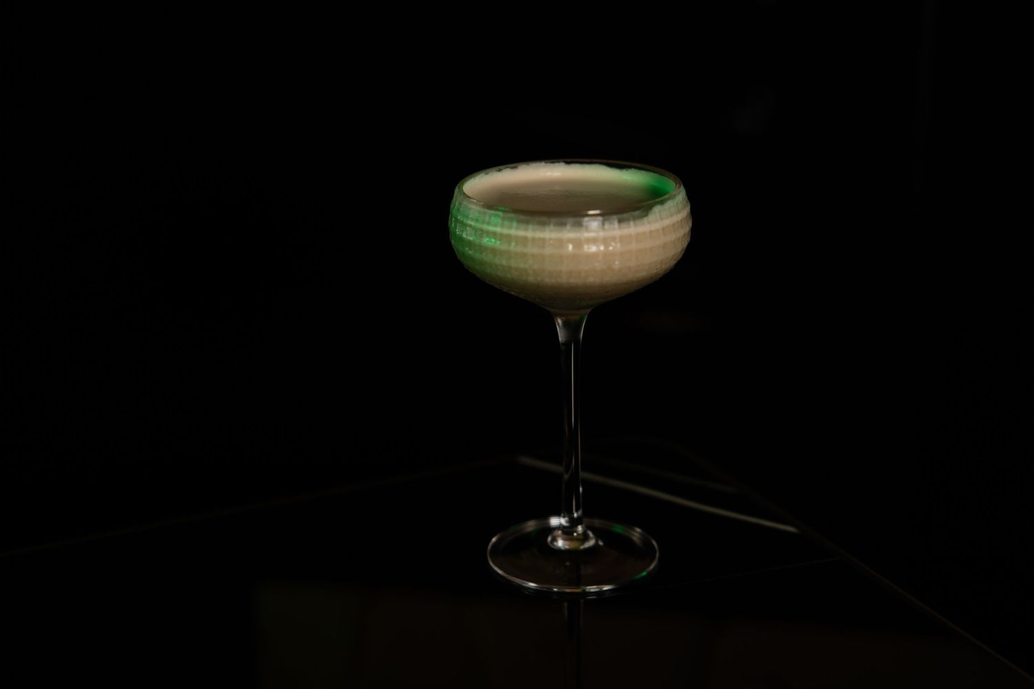 Elegant coupe glass filled with a dark cocktail topped with green foam against a black background.