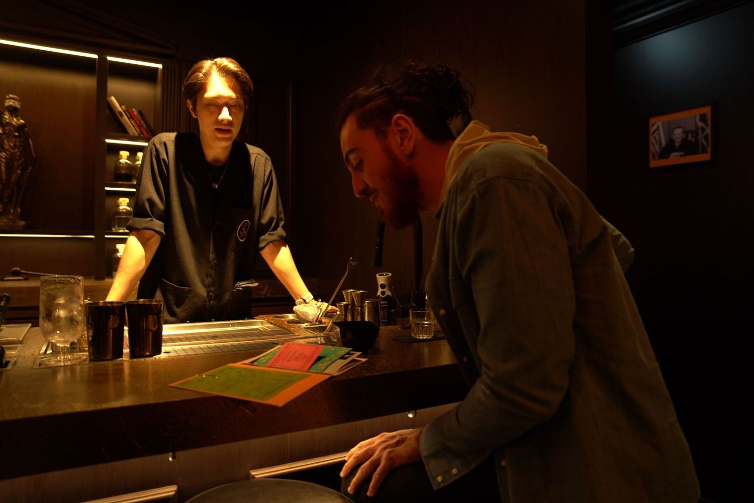 Two people interact across a dark wooden bar counter in a moody nighttime setting.