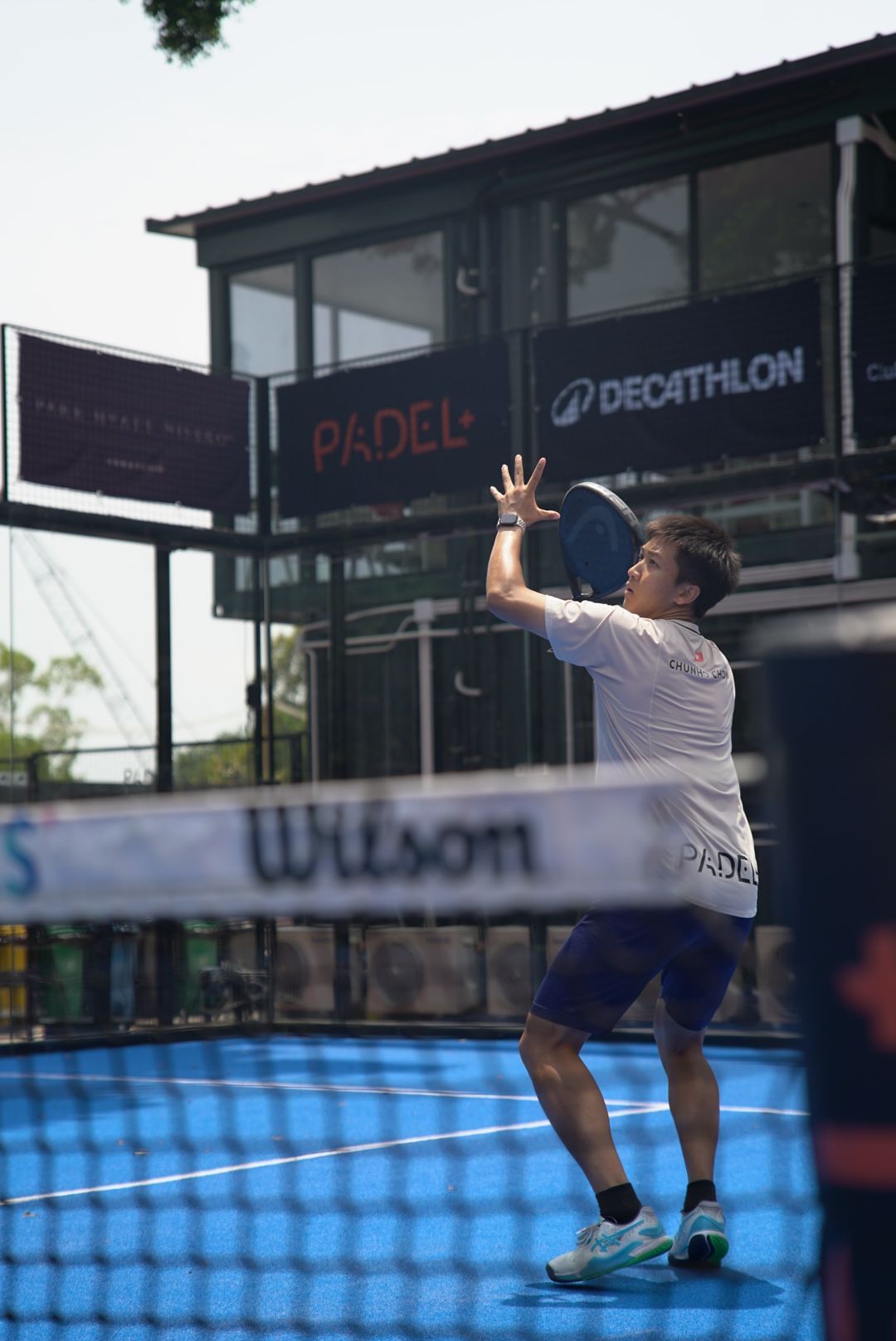 Player serving during padel match at Decathlon court with blue floor and Wilson net in background.