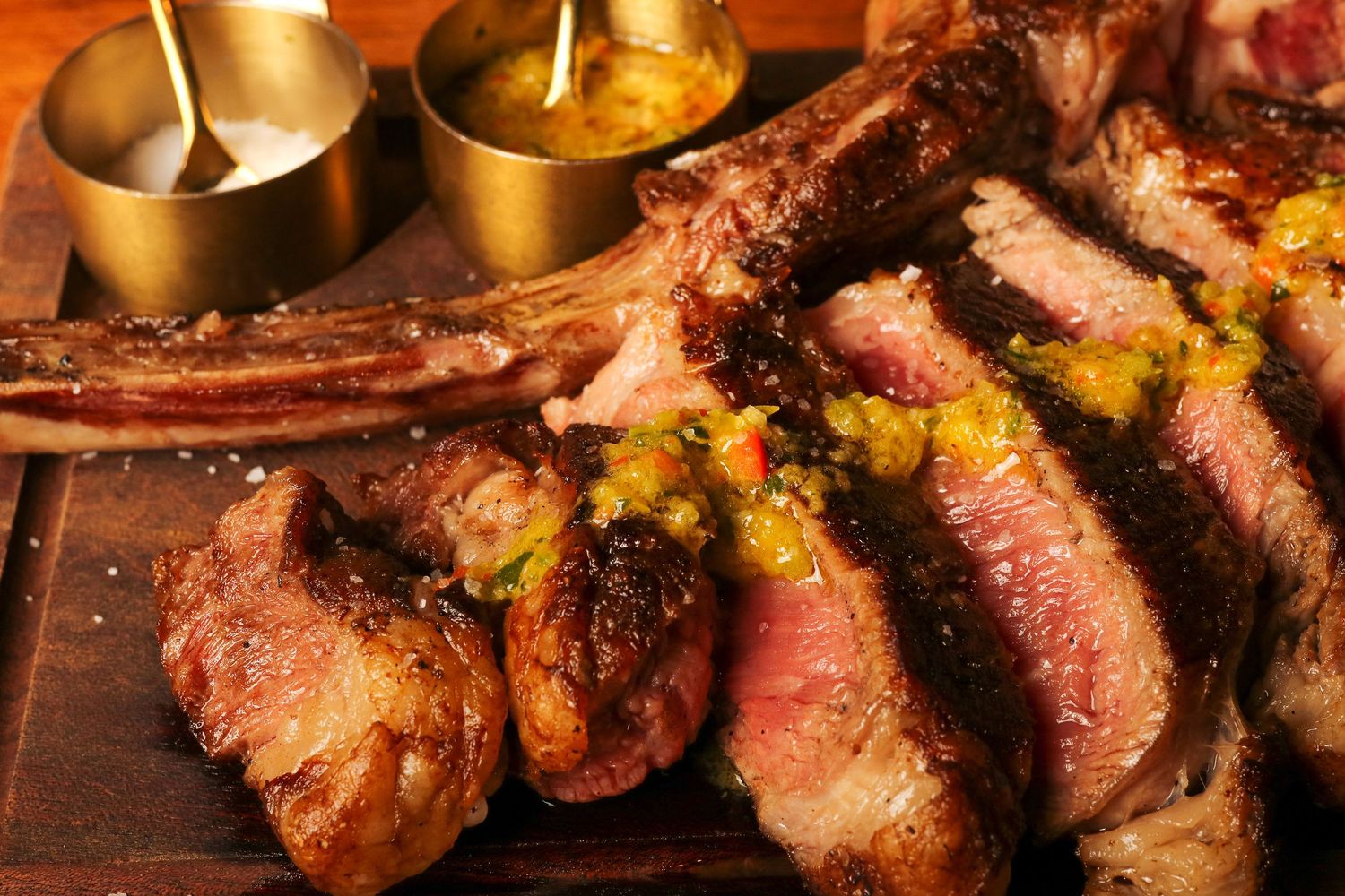 Juicy sliced medium-rare tomahawk steak on wooden board with sauce bowls in golden lighting.