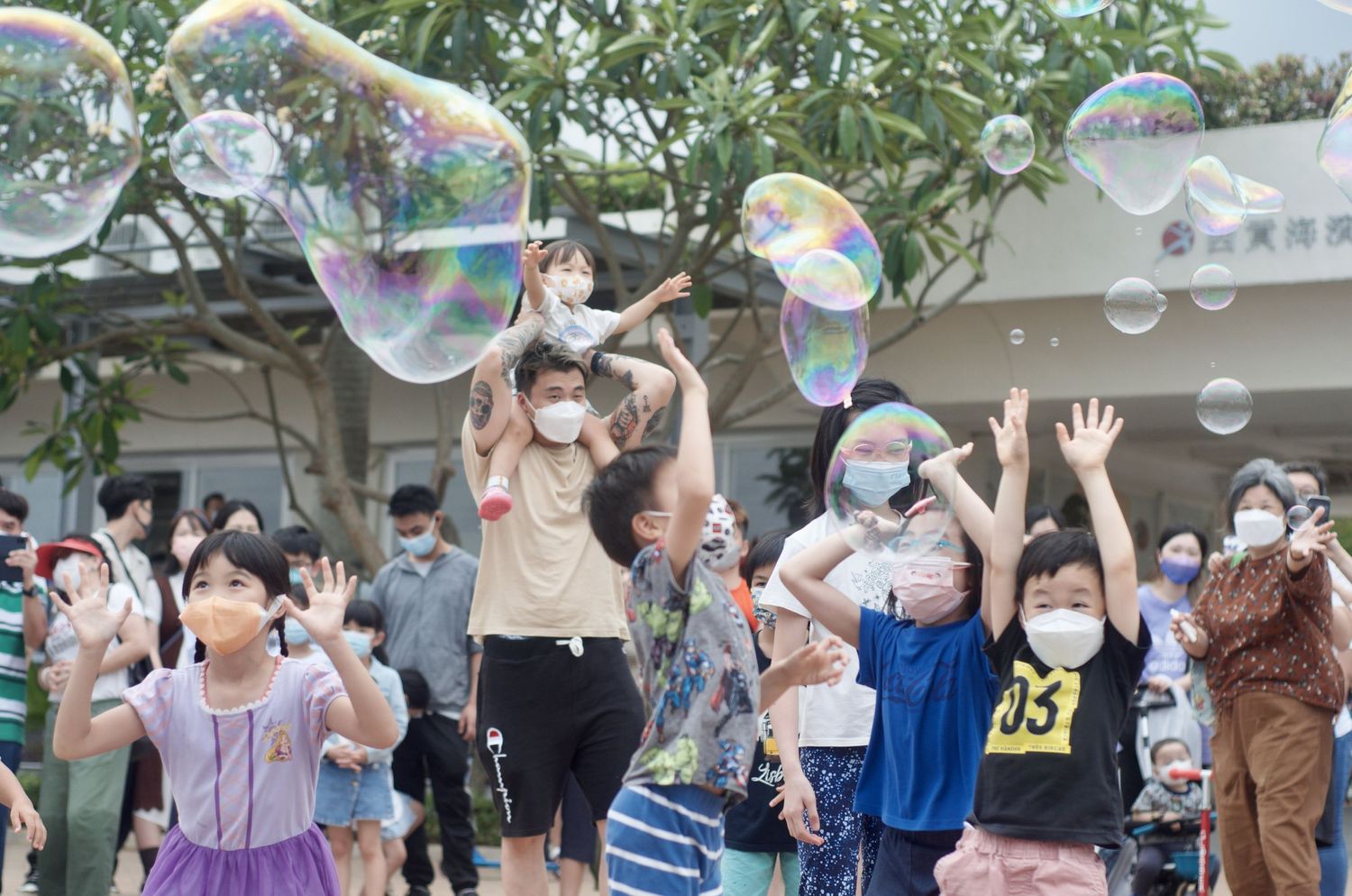 Crowd of people reaching up to catch giant soap bubbles against a tree backdrop.