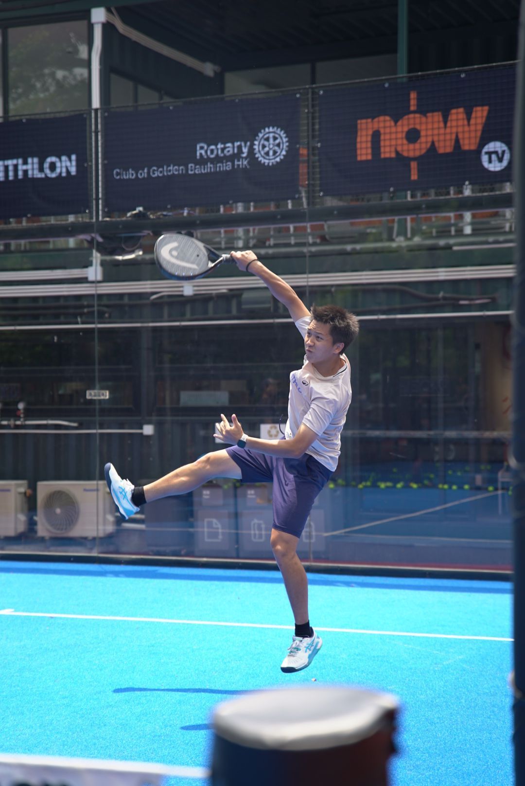A padel tennis player jumps high to hit an overhead smash on a blue court during a match.