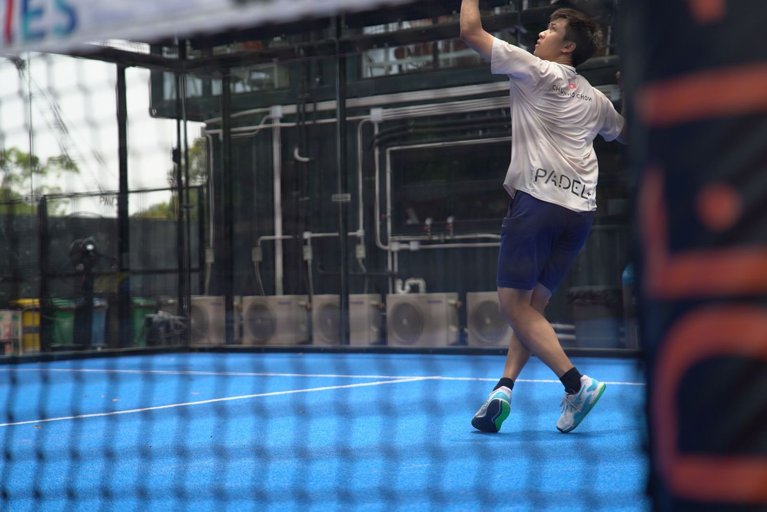 A player leaps high to make an overhead shot during a padel tennis match through glass enclosure.
