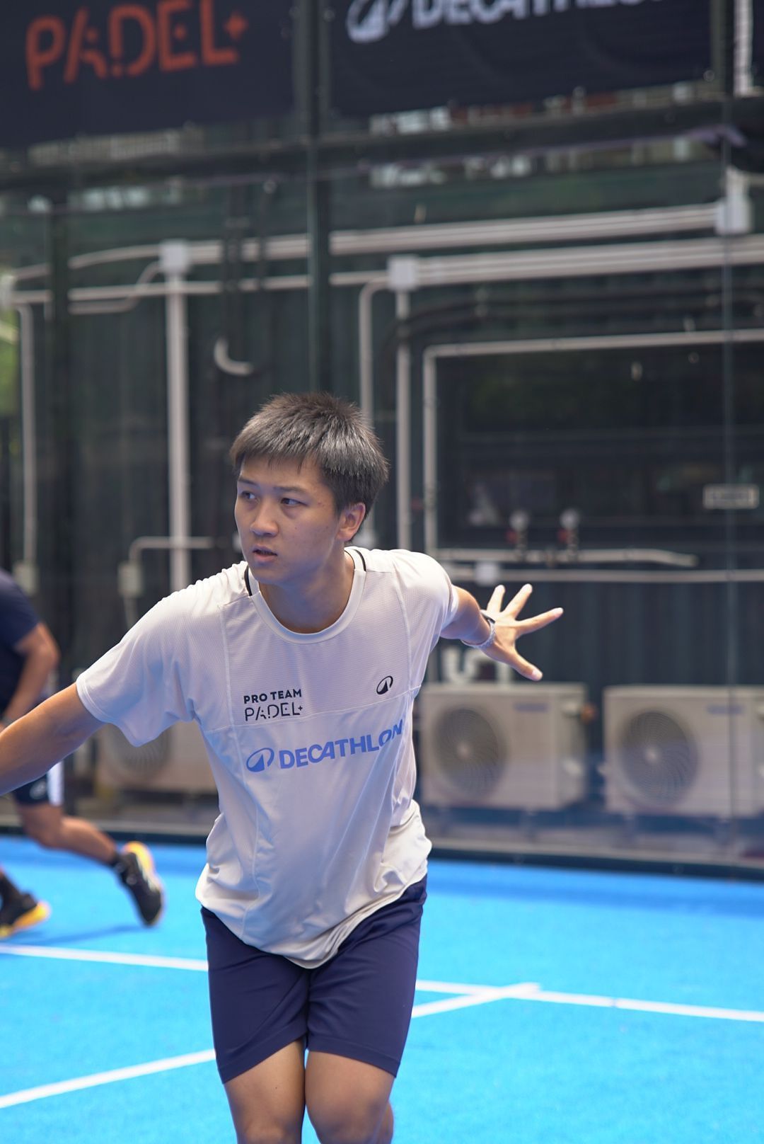 Player wearing Decathlon white t-shirt preparing to serve during padel match on blue court.
