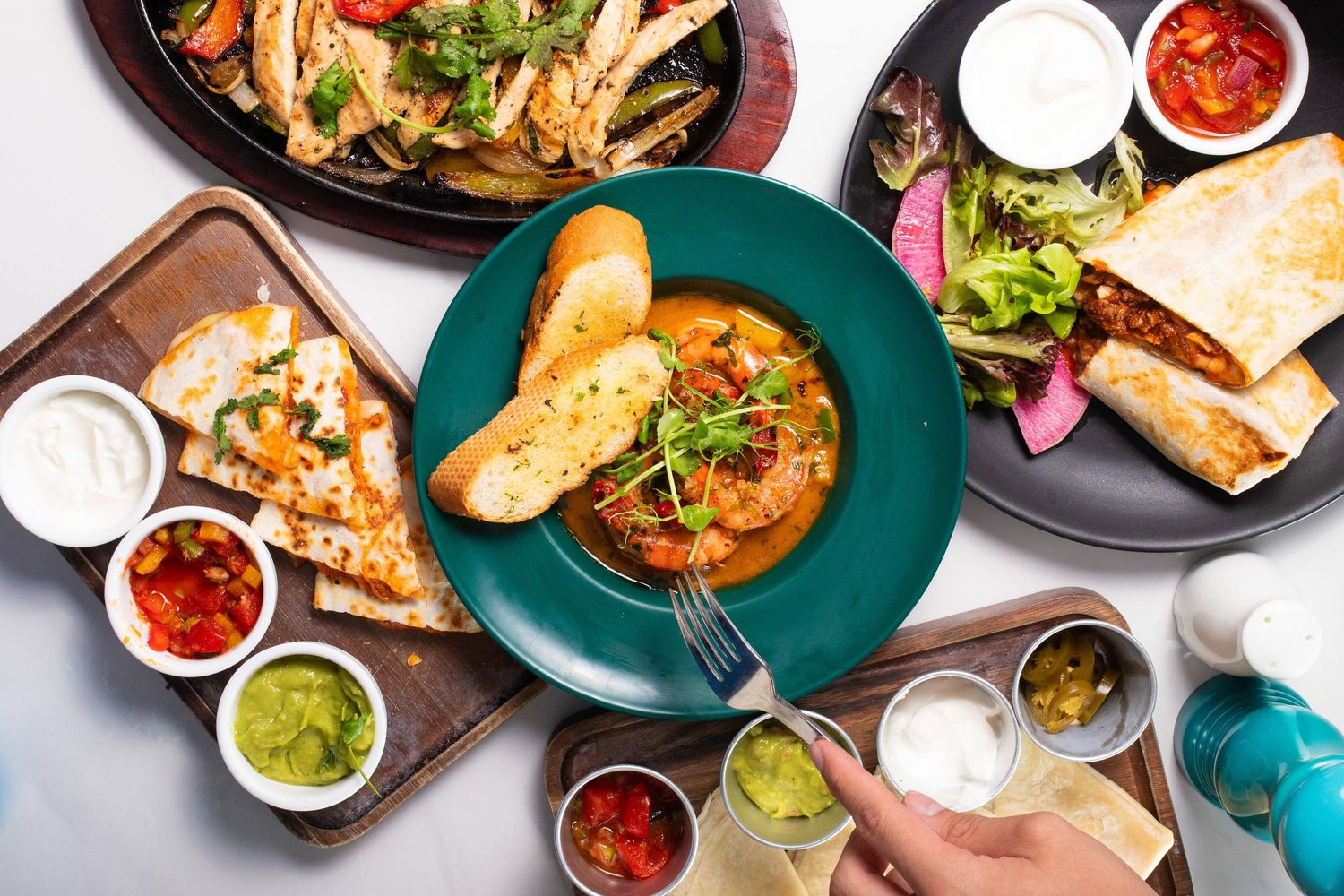 Overhead view of Mexican restaurant table with spicy soup, tortas, salsas, and sides on colorful plates.