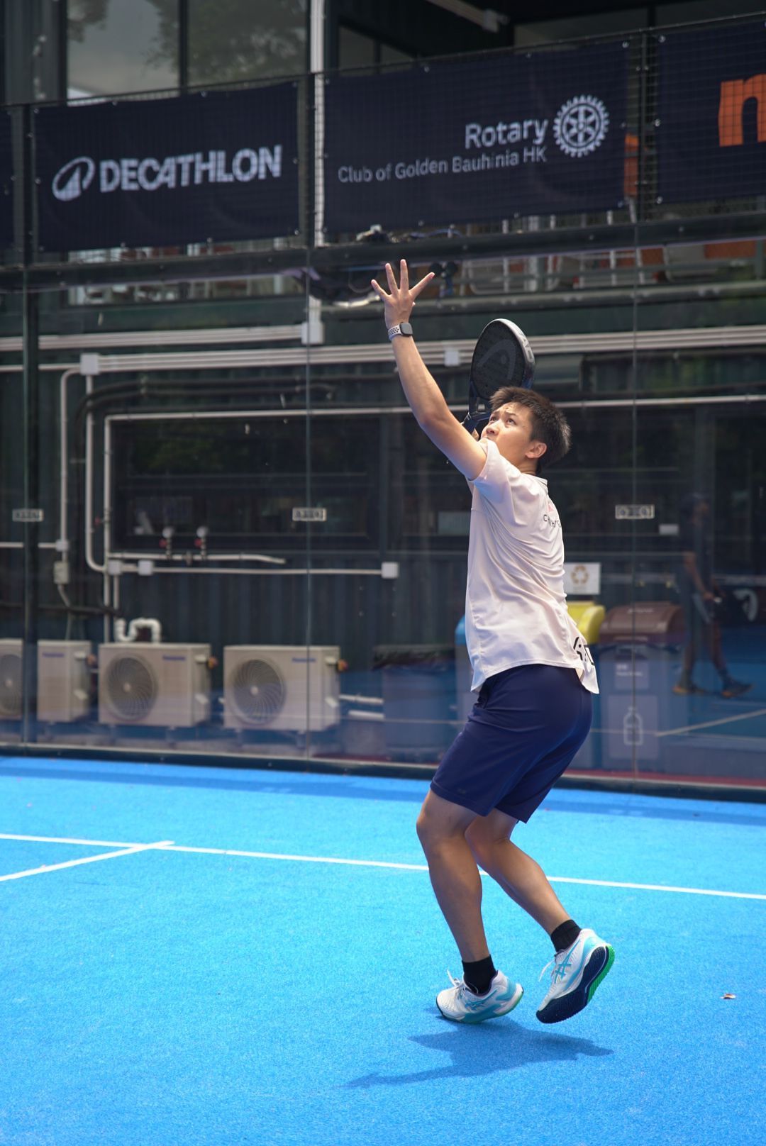 Player serves on blue padel court outside Decathlon store with Rotary Club signage visible in background.