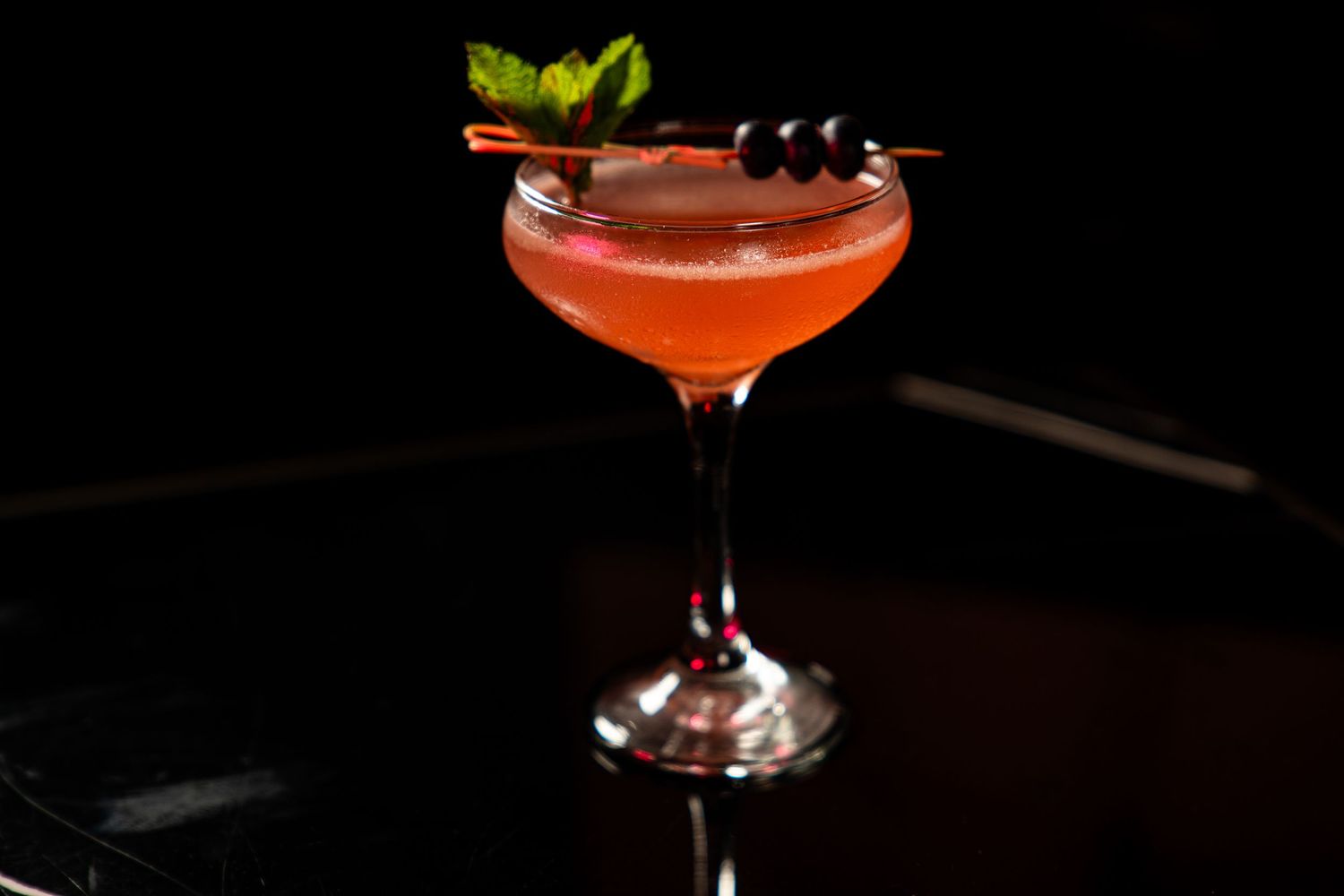 A red cocktail garnished with black berries and green herbs served in a coupe glass against a dark background.