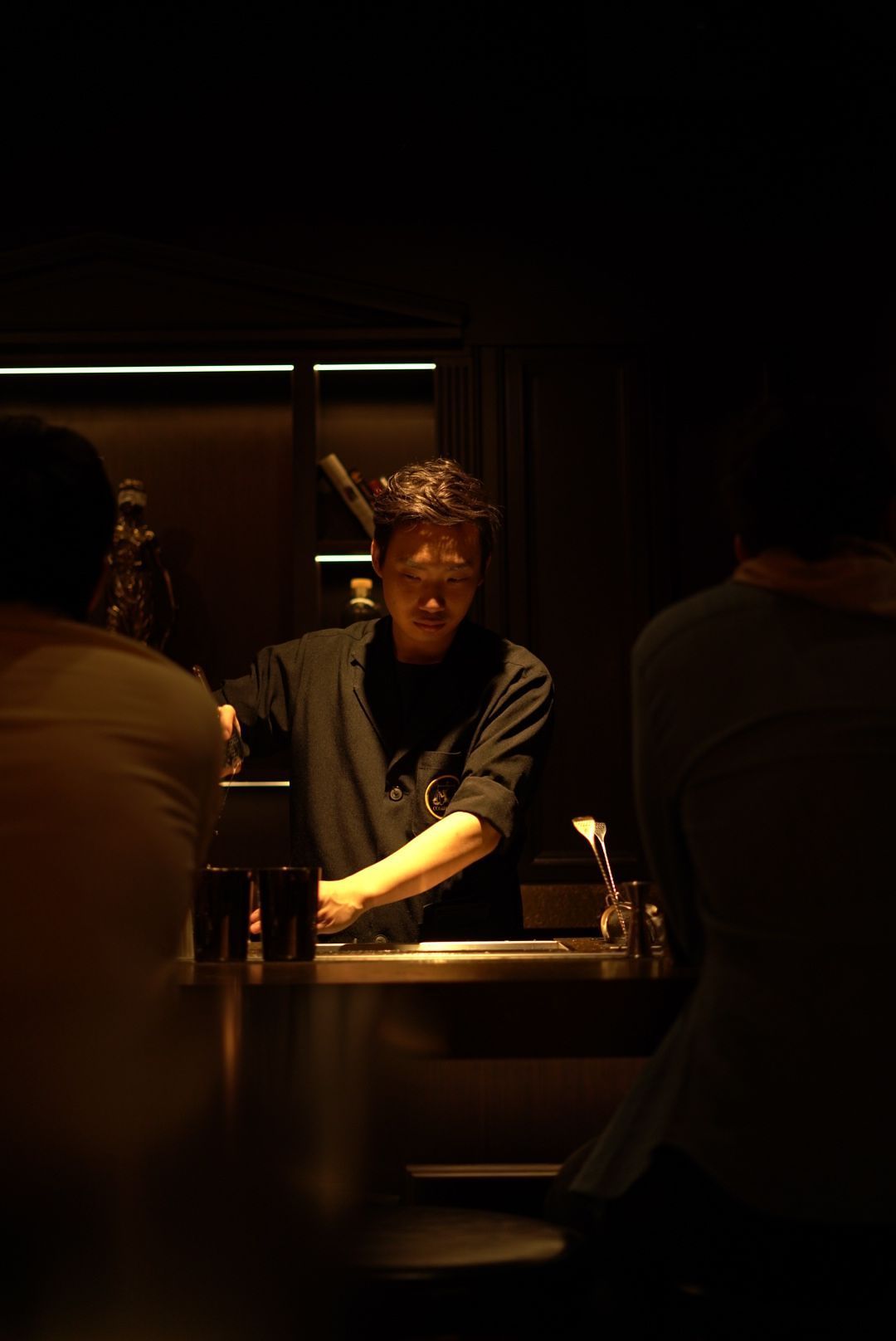 A bartender works behind a dimly lit bar counter, illuminated by soft overhead lighting while mixing drinks.