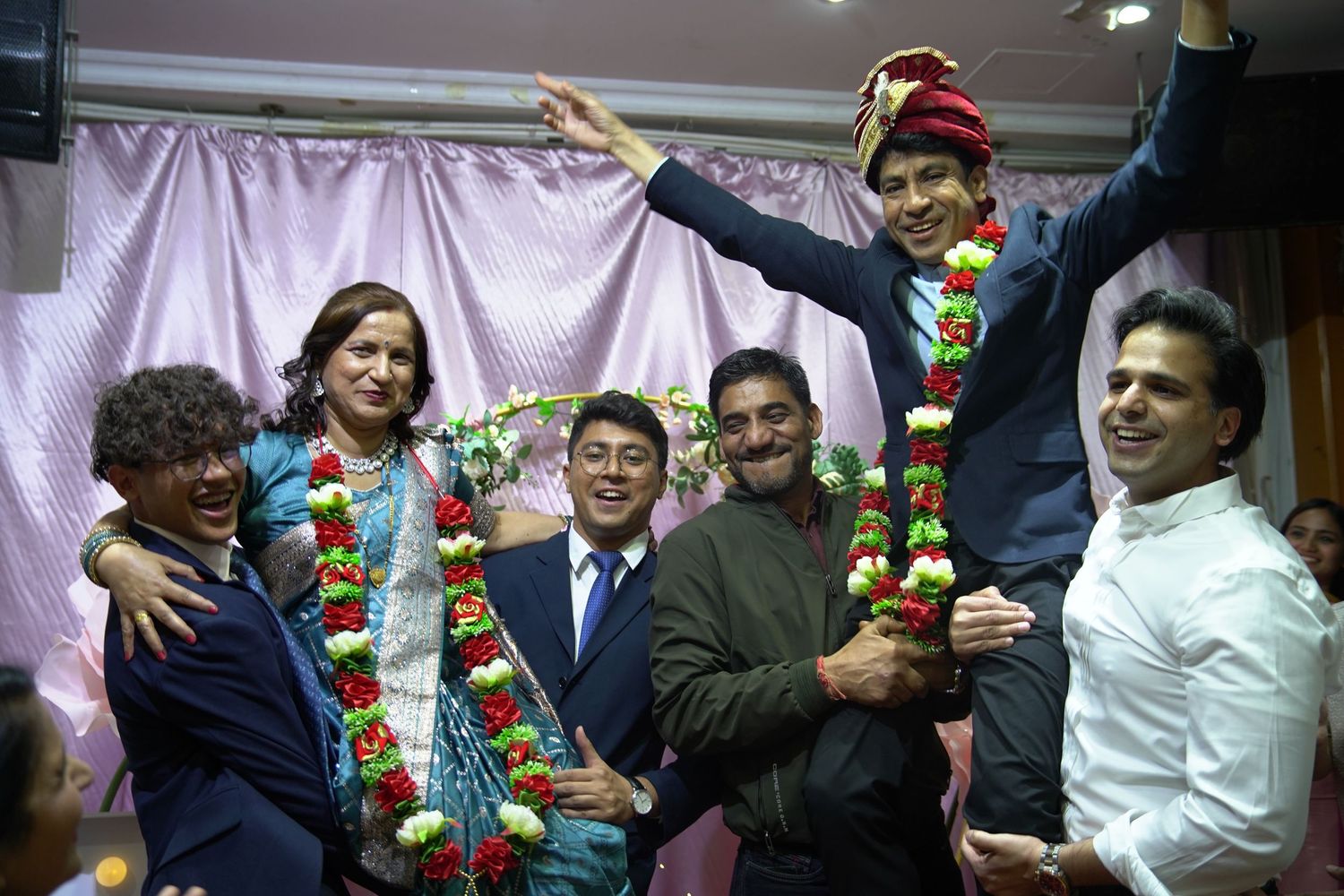 A group celebrates in a festive setting with colorful garlands and decorative ornaments at an indoor gathering.