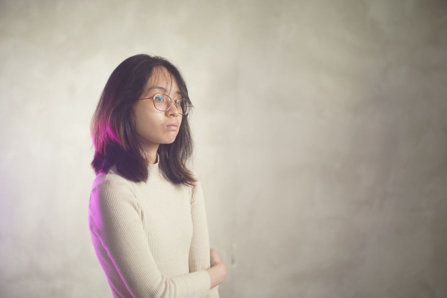 Person in white sweater stands thoughtfully against textured wall with purple lighting effect in moody portrait.