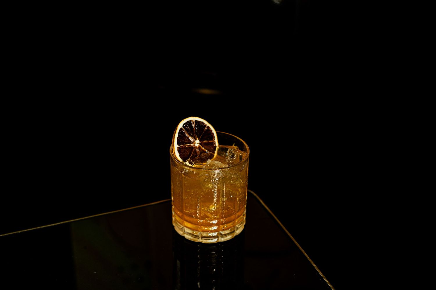 A golden cocktail garnished with a citrus wheel sits elegantly on a dark surface in dim lighting.