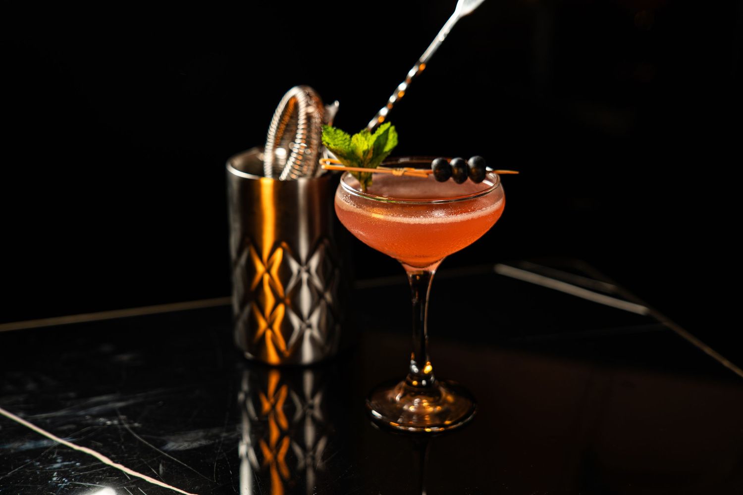 Two elegant cocktails on a dark bar counter, one in a copper patterned tumbler and one in a coupe glass garnished with herbs.