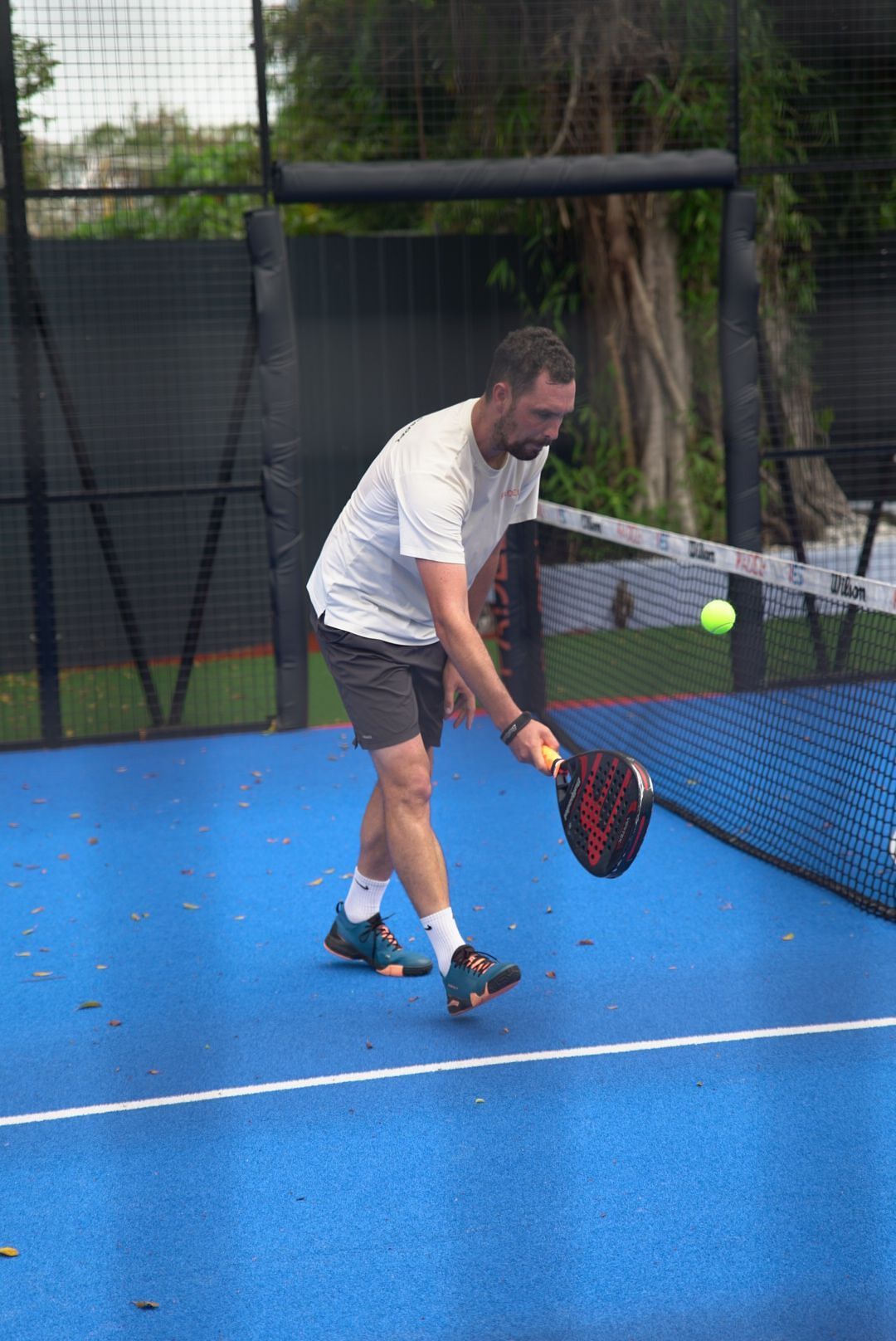 Person playing paddle tennis on outdoor blue court hitting yellow ball with red racquet during daytime sport activity.