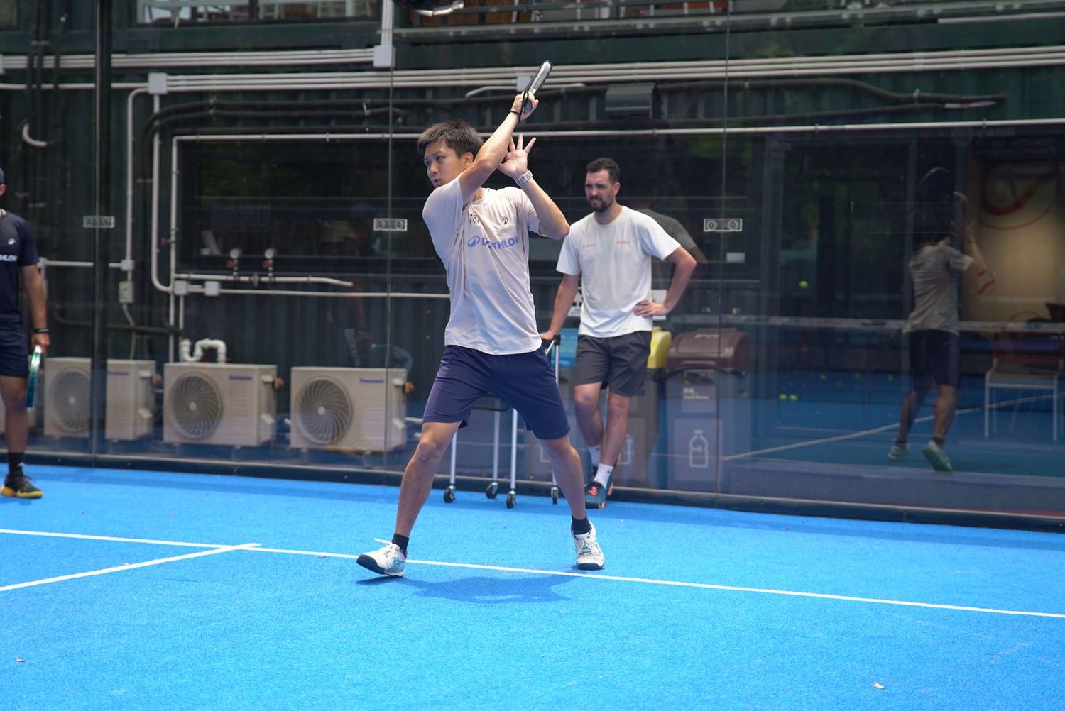Players practicing on an outdoor blue padel court with protective glass walls and fencing on a sunny day.
