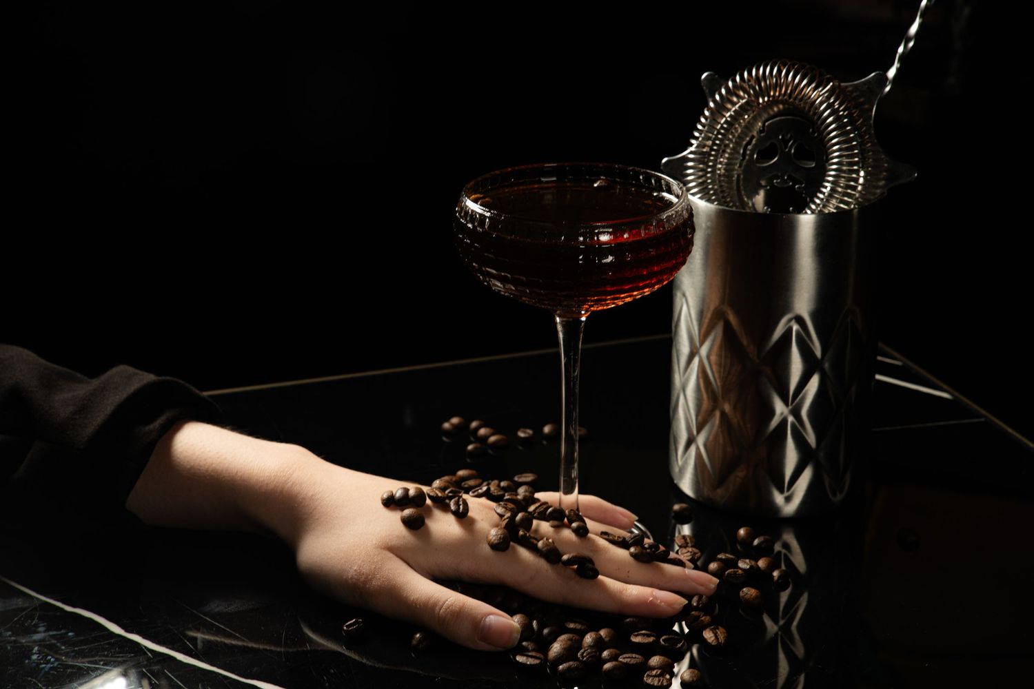 Coffee beans spilling from ceramic hand decor with cocktail glass and metal shaker against dark background.