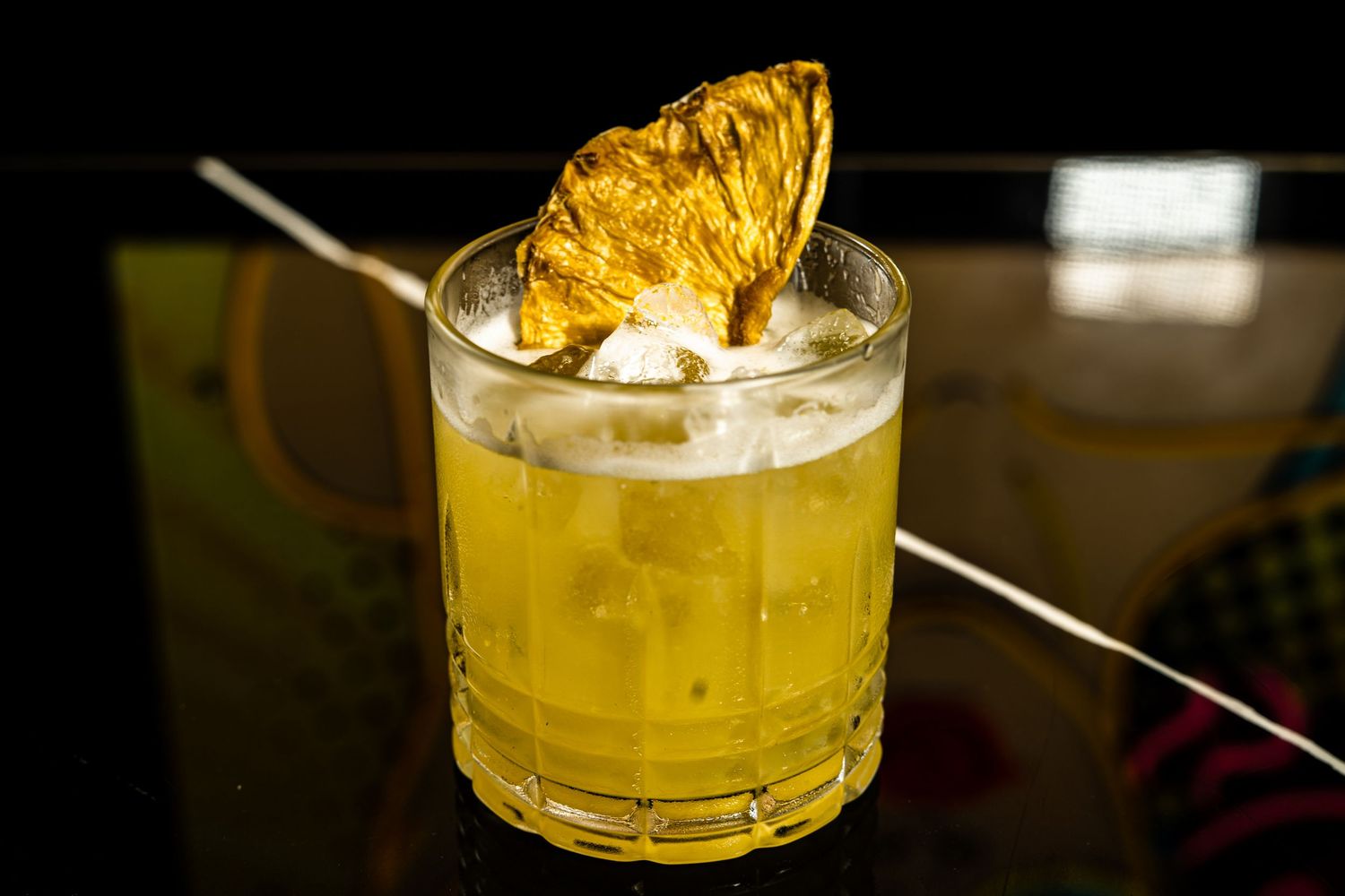 Yellow cocktail garnished with dried pineapple in crystal rocks glass on dark surface.