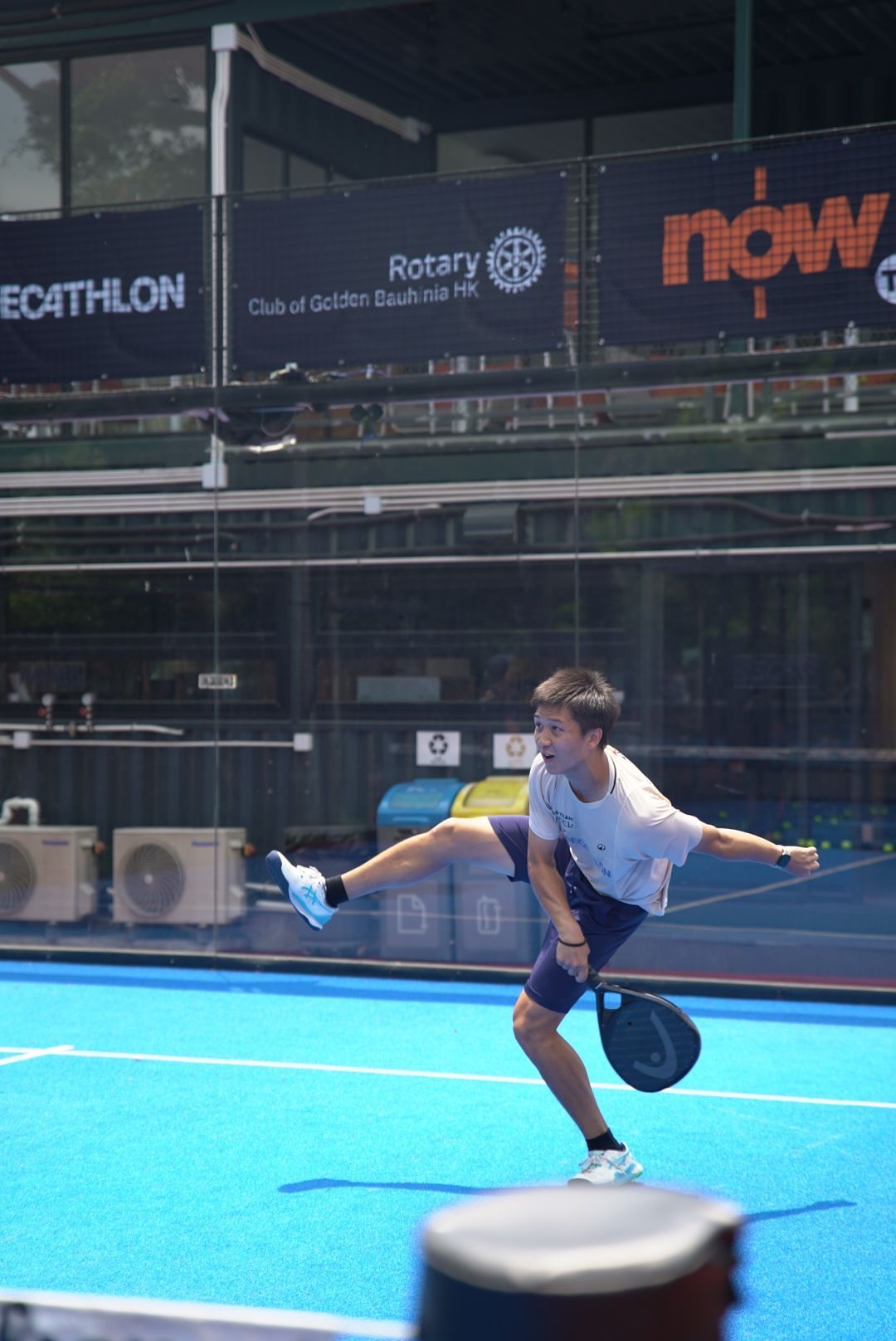 Tennis player executing a dynamic serve motion on a blue hard court with Decathlon and Rotary Club signage visible.